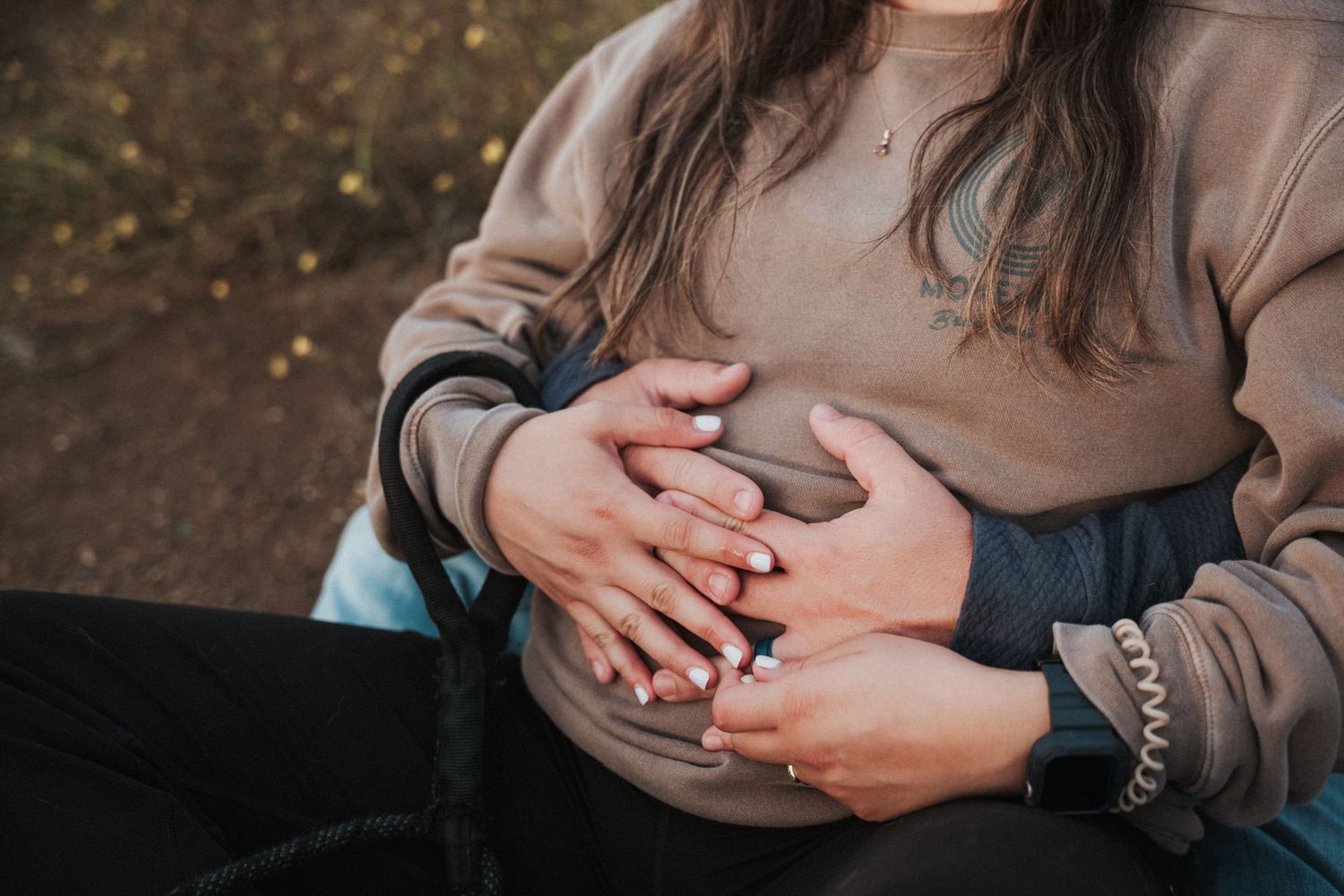 Couple tenderly holding pregnant belly outdoors, wearing casual clothing in a warm autumn setting.