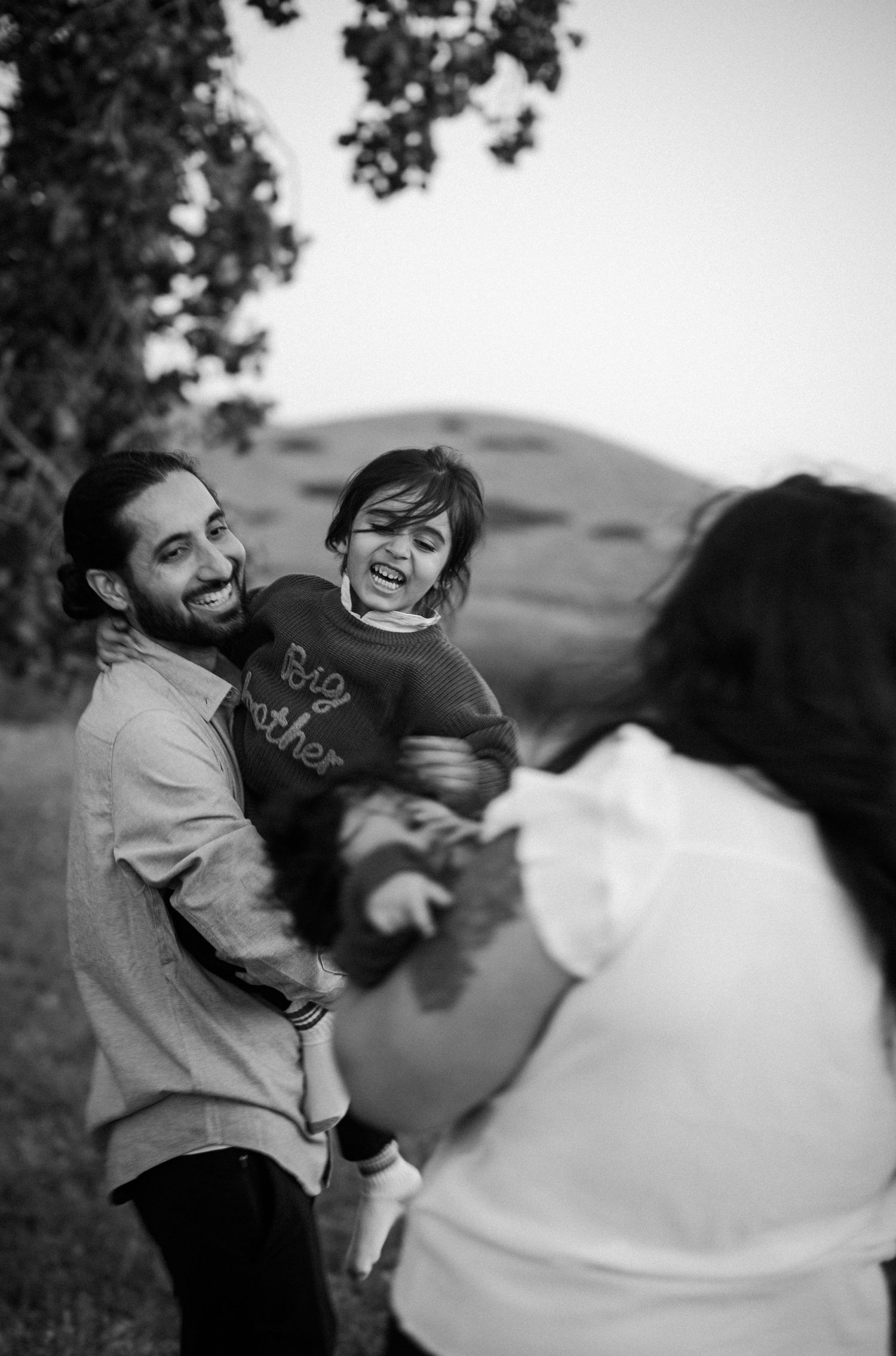 Black and white image of smiling father holding giggling child outdoors with rolling hills in background.