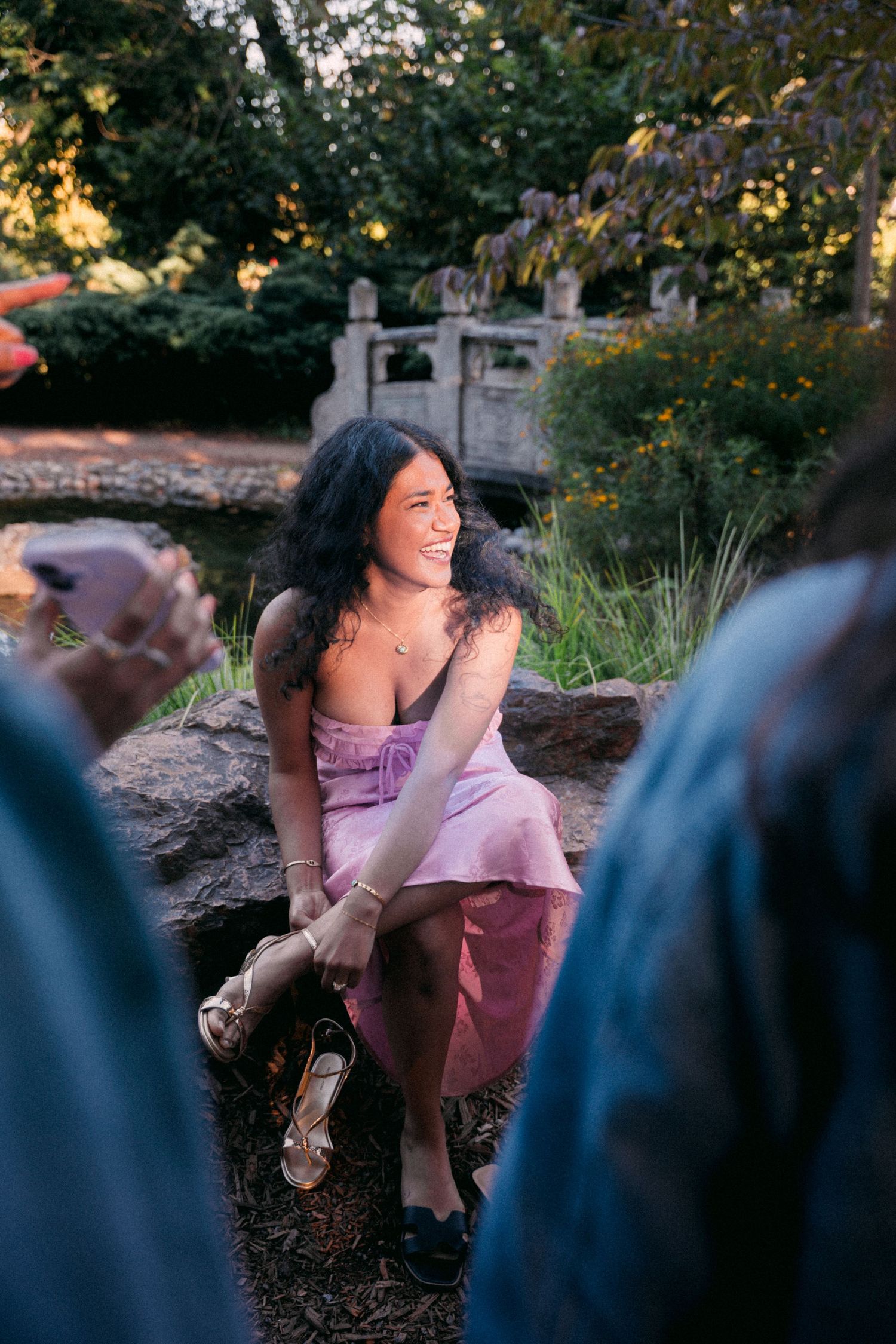 A smiling woman in a pink dress sits on rocks in a lush garden near a stone bridge at sunset.