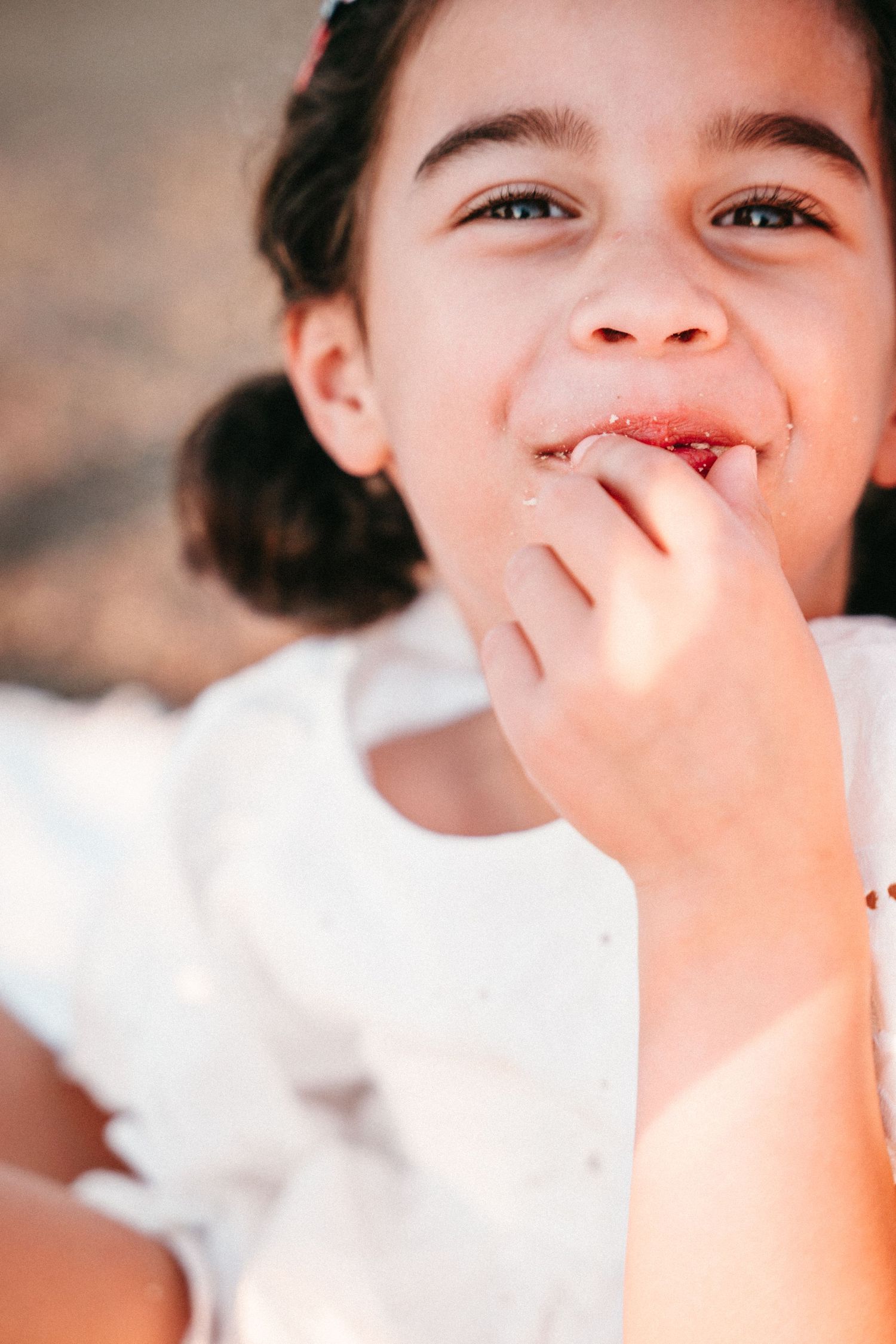 A young girl in a white dress smiles while licking her fingers outdoors in natural sunlight.