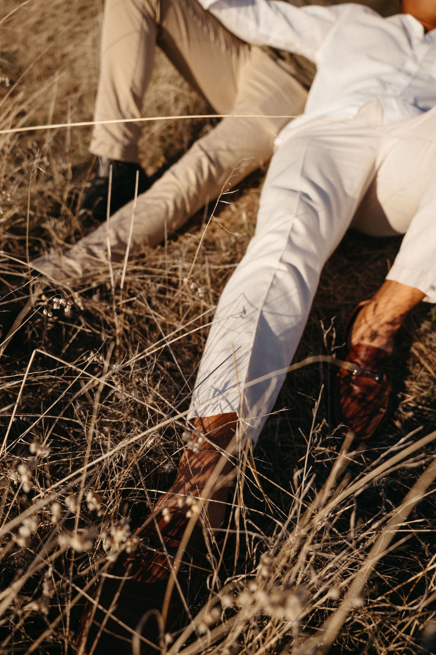 Two people sitting in dry grass field wearing white and beige outfits with brown leather shoes on a sunny day.