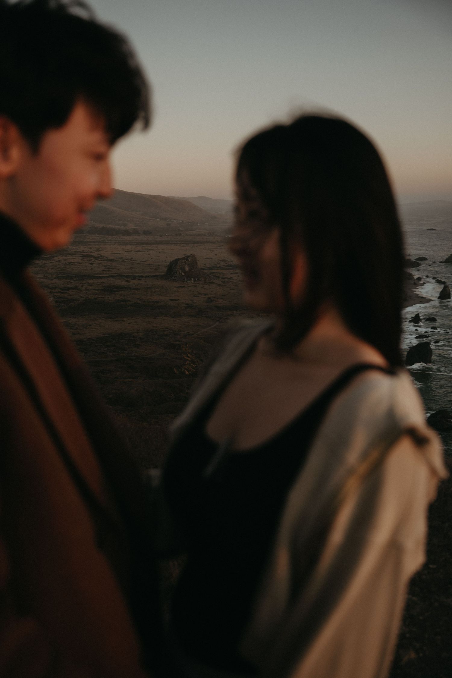 A softly blurred couple stands face to face on a coastal bluff at dusk, with the ocean and rolling hills behind them.