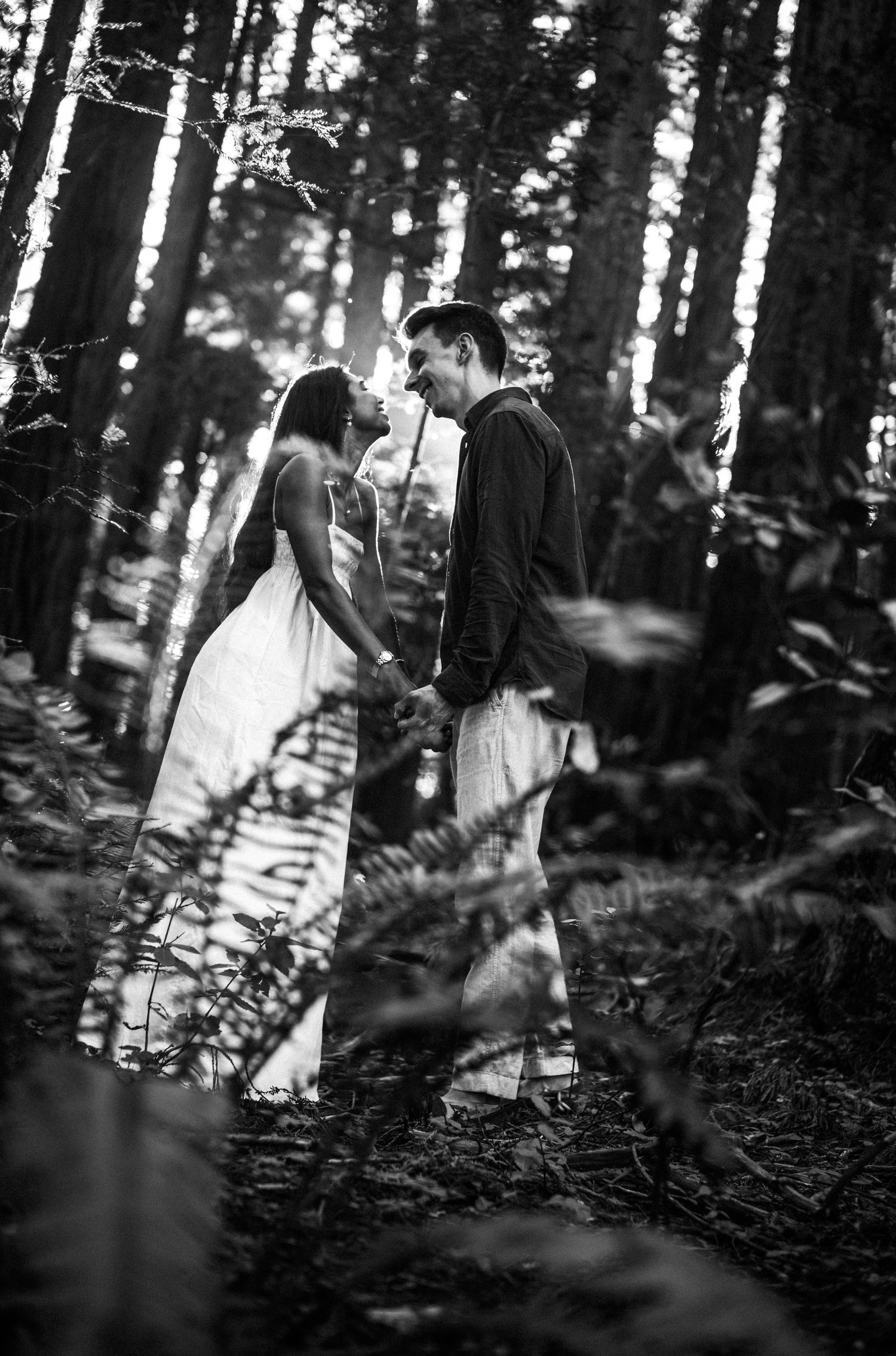 Black and white photo of a couple holding hands and smiling at each other in a lush forest setting.