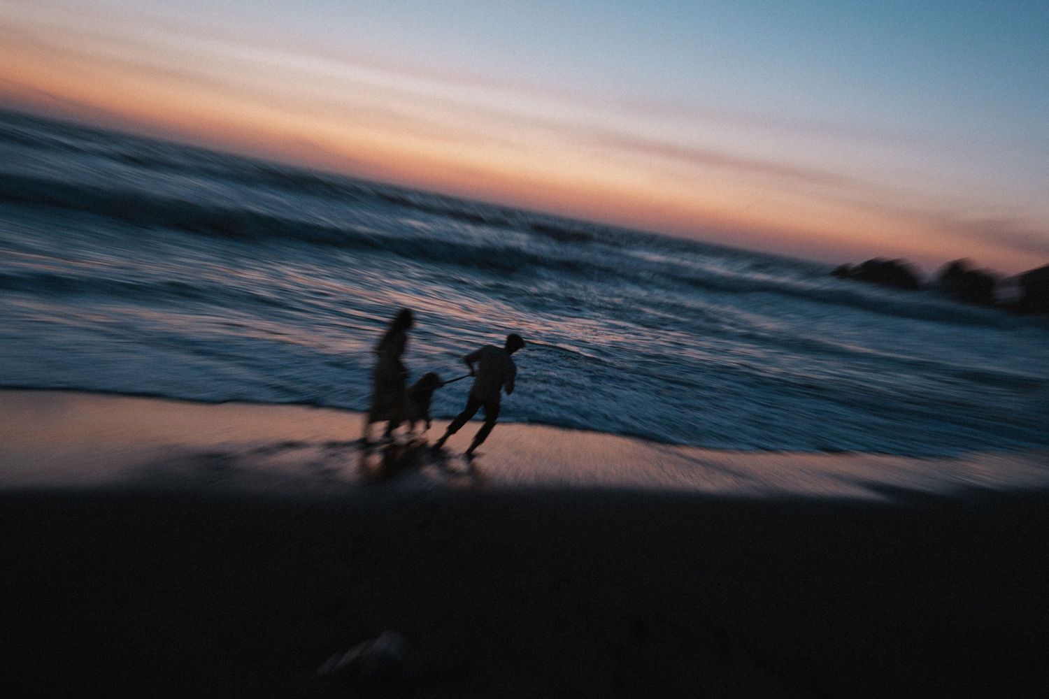 Silhouette of couple running with dog along beach shoreline at dusk with colorful sunset sky.