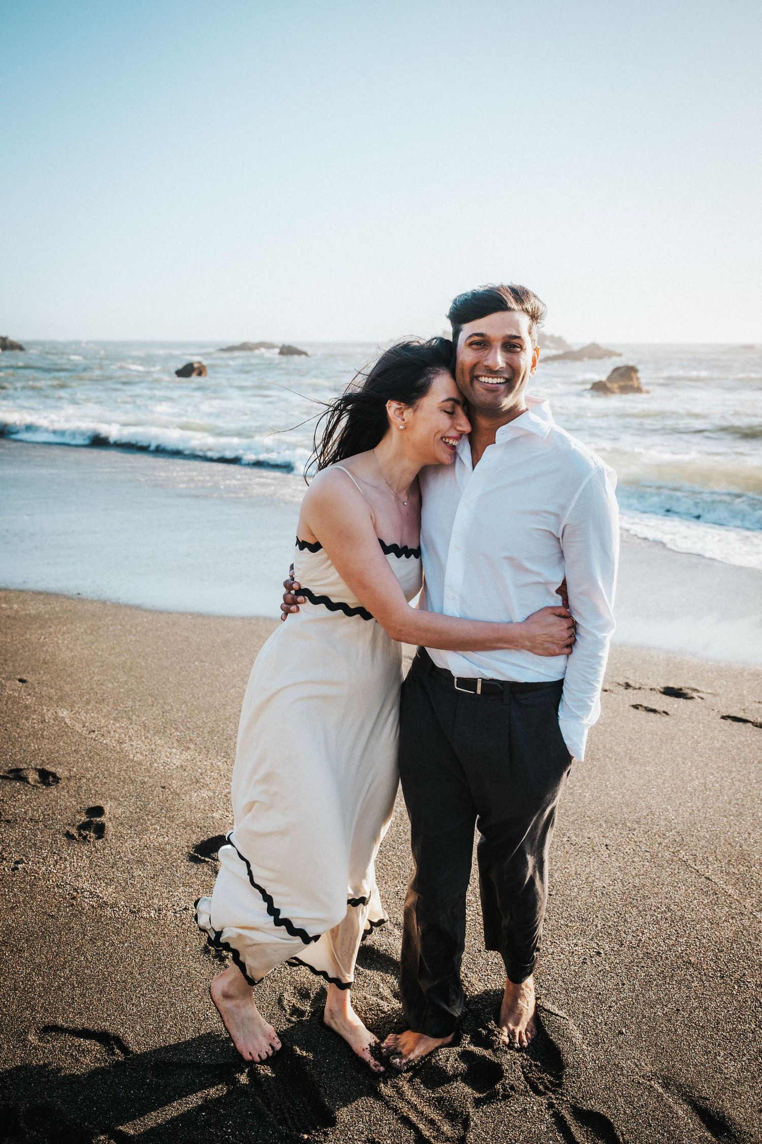 A smiling couple embraces barefoot on a sandy beach with ocean waves, woman in white dress, man in white shirt and gray pants.