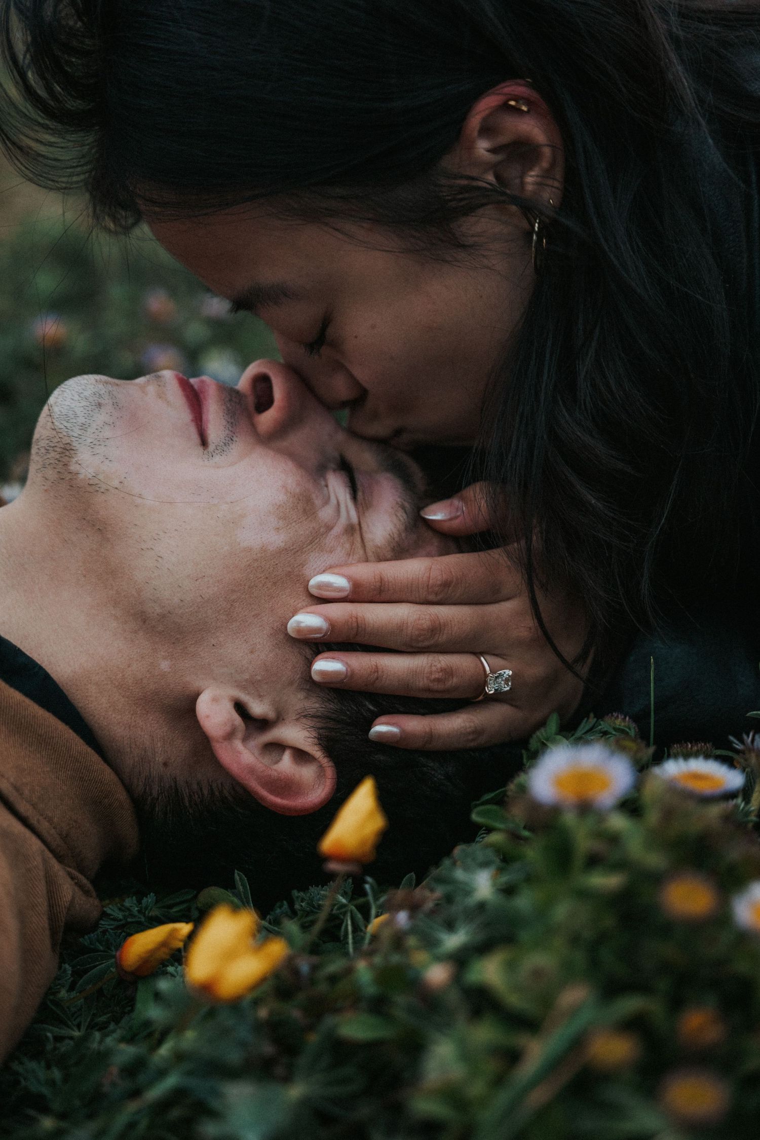 Couple sharing a romantic kiss outdoors among wildflowers, woman wearing engagement ring cradling mans face.