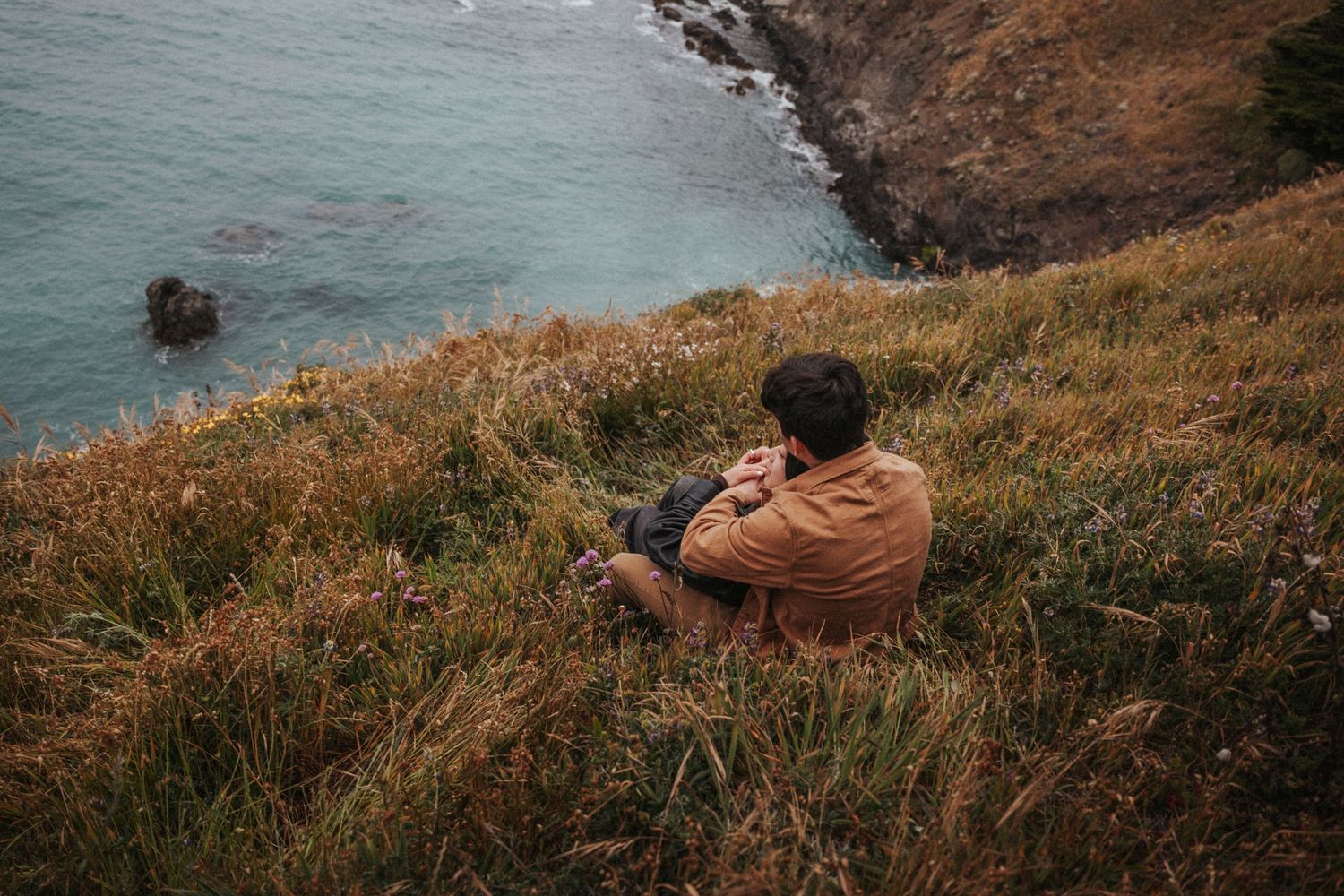 Man in brown jacket sits on grassy coastal cliff overlooking rocky ocean shoreline.