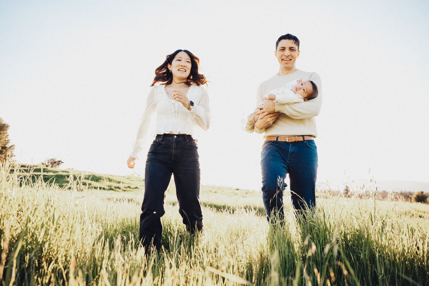 Young family with newborn baby stands in a sunlit green field, enjoying a peaceful outdoor moment.