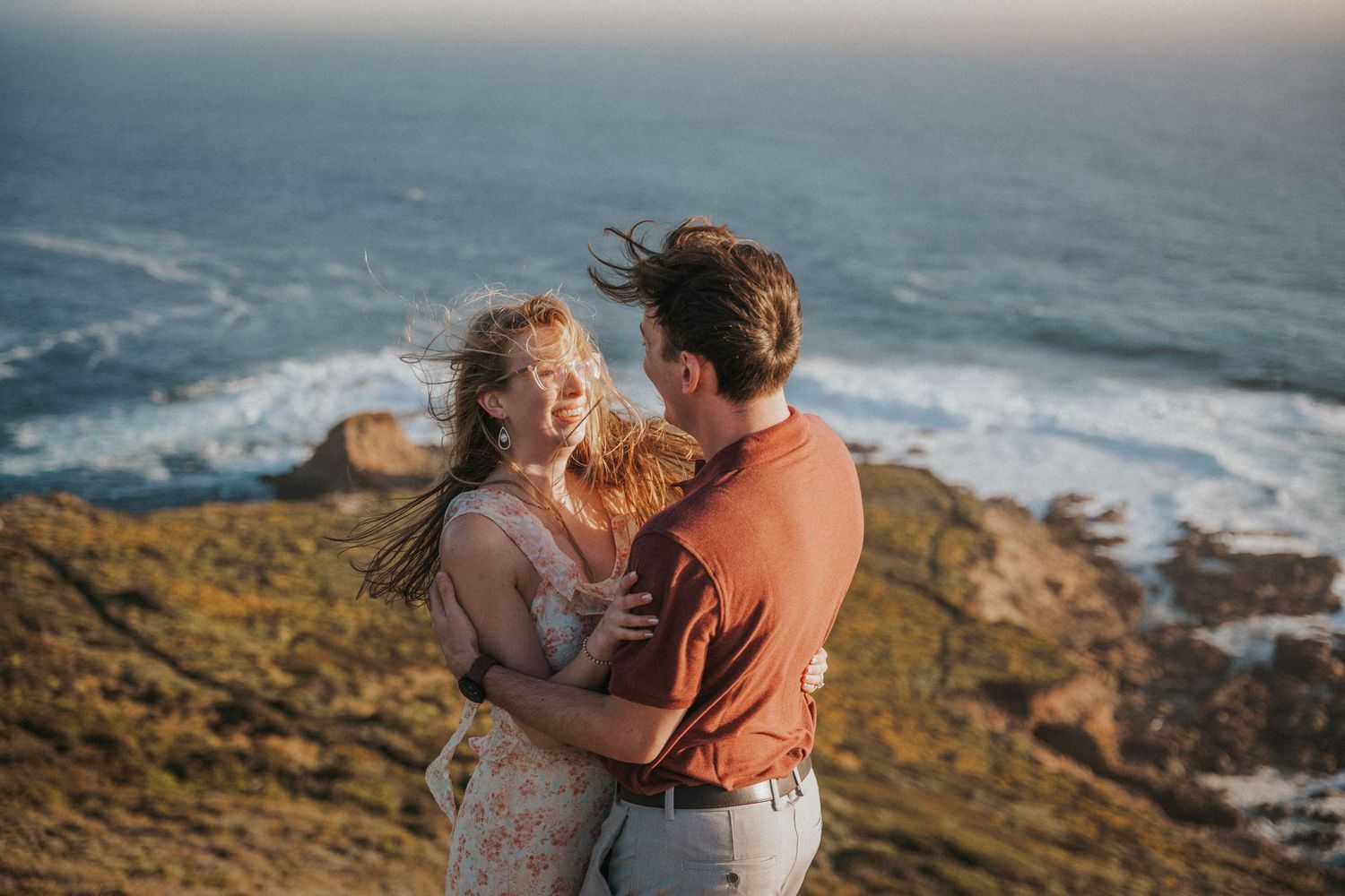 Couple embracing on a coastal cliff at golden hour with ocean waves behind them.