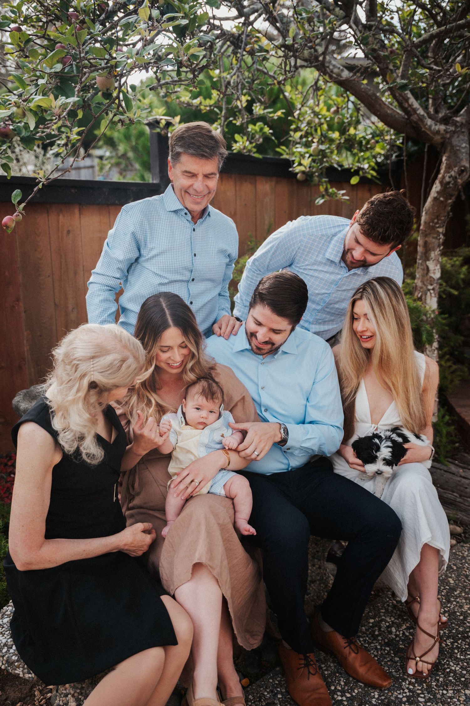 Multi-generational family portrait outdoors, gathering around a baby, with a puppy, smiling warmly beneath a tree.