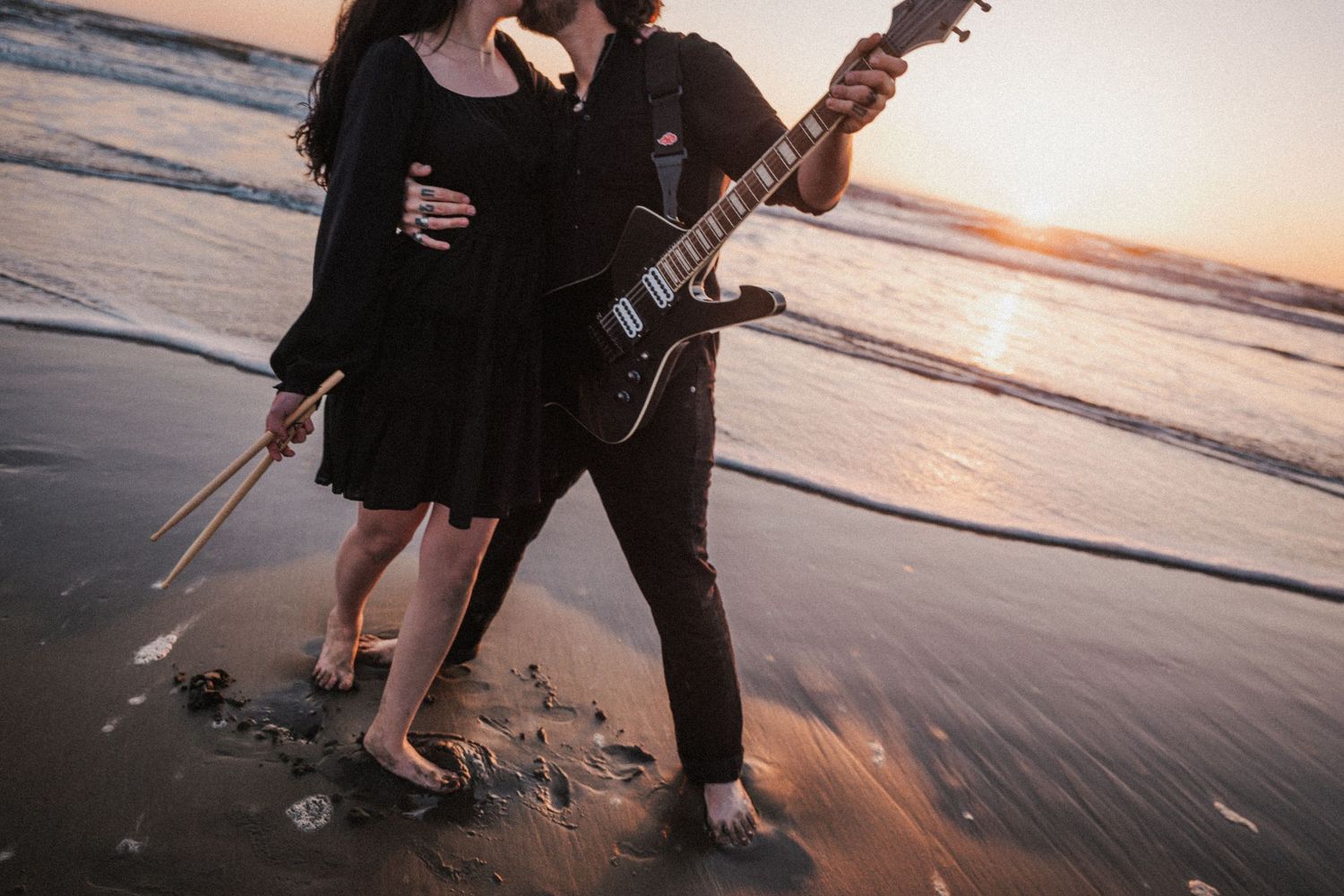 Barefoot couple on beach at sunset, woman holding drumsticks, man playing black electric guitar.
