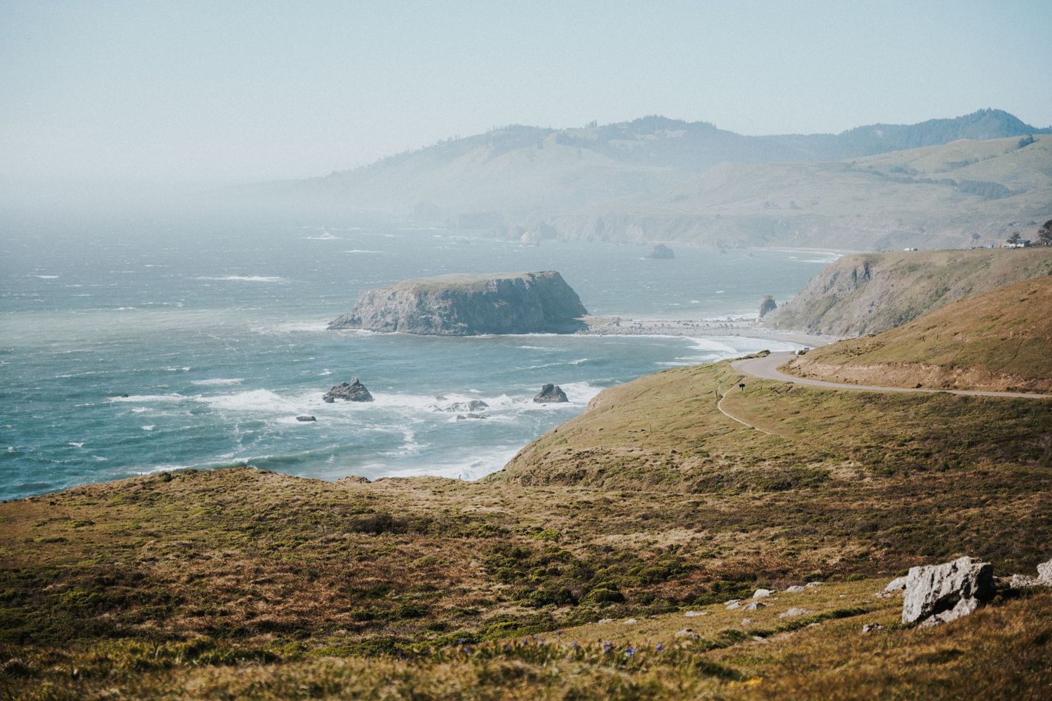 Scenic coastal vista featuring rocky sea stacks, rolling green hills, and a misty mountain backdrop by the ocean.