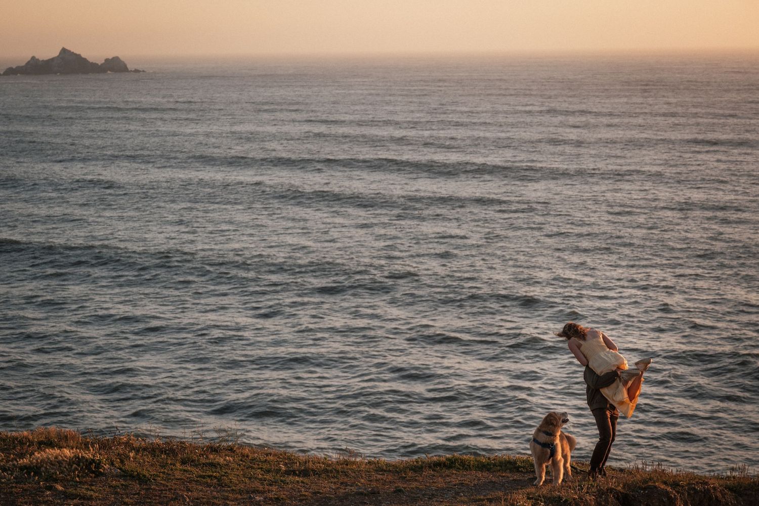 Person and dog standing on coastal cliffside at golden hour sunset with ocean and rocky island in background.