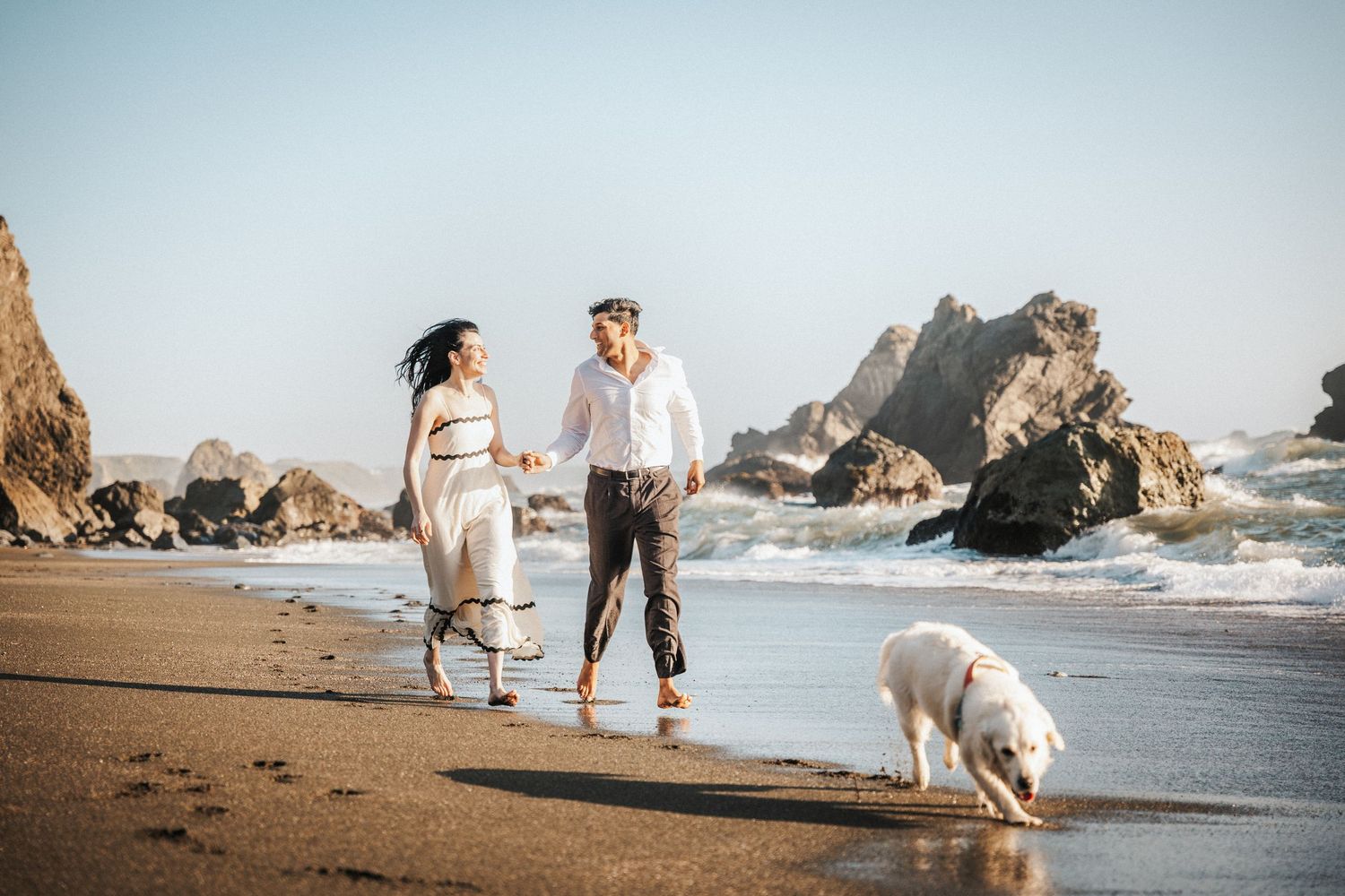 Couple walking barefoot on sandy beach with white dog, woman in white dress, man in white shirt, ocean waves behind.