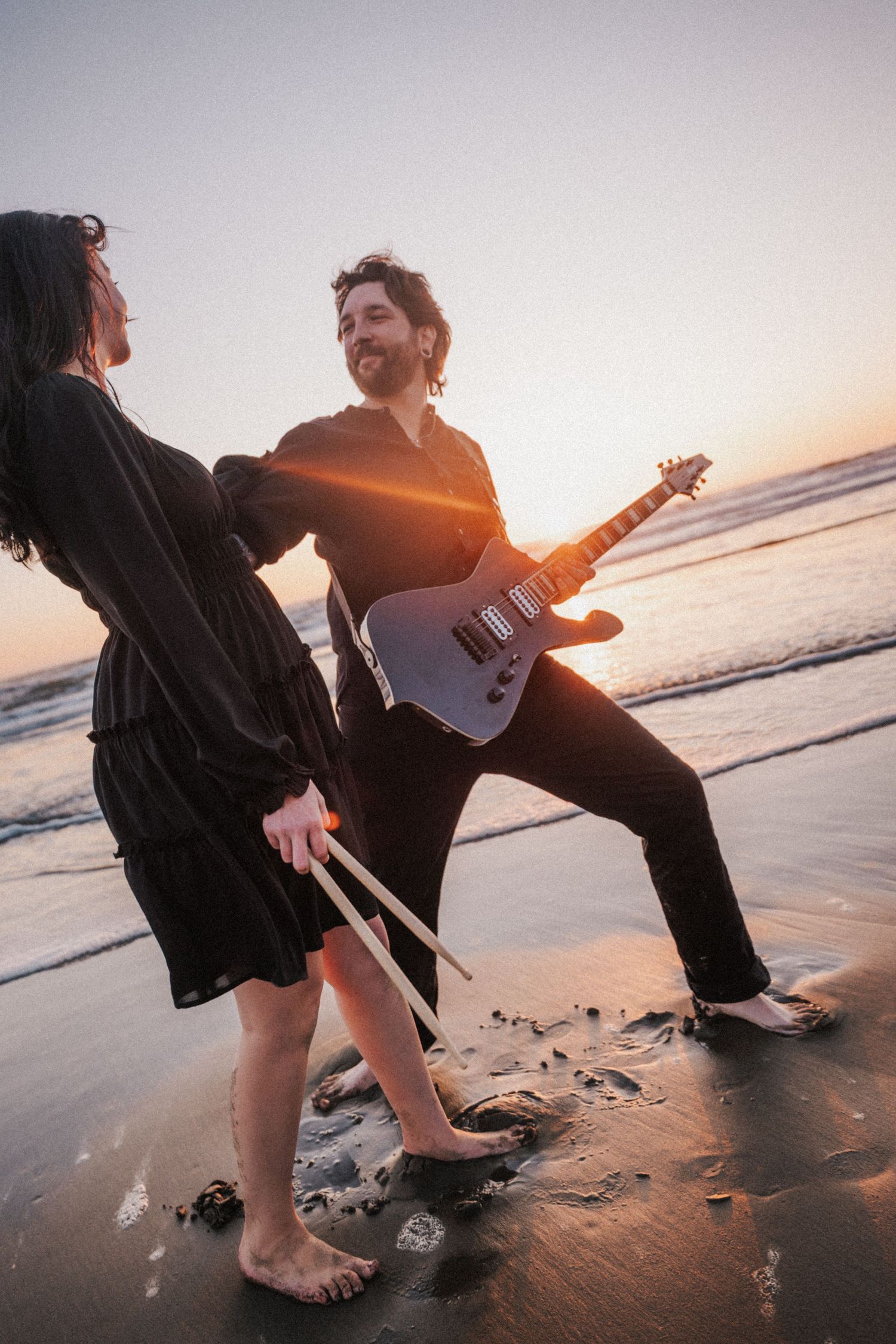 Couple dancing on beach at sunset, man playing electric guitar while woman holds drumsticks.