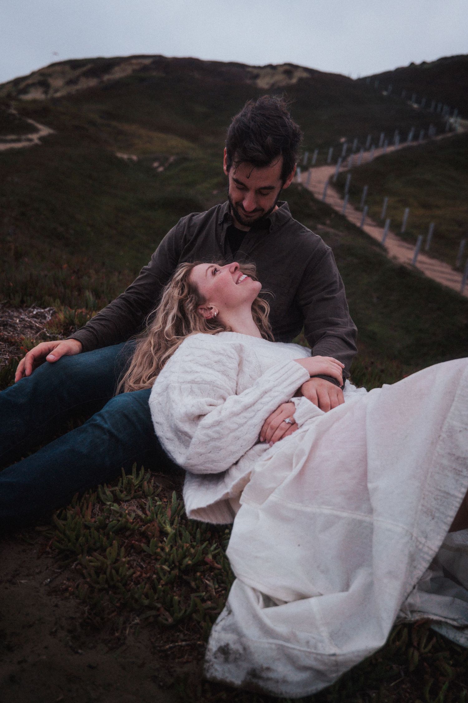 A couple relaxes on a green hillside, the woman lying in the man's lap, smiling up at him.