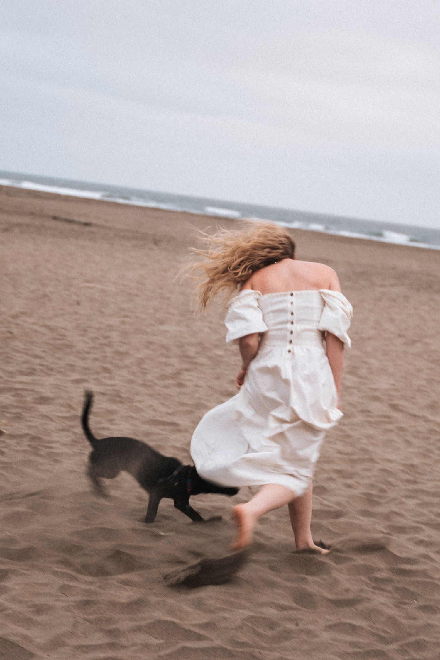 Woman in white off-shoulder dress running barefoot on beach with black dog on overcast day.