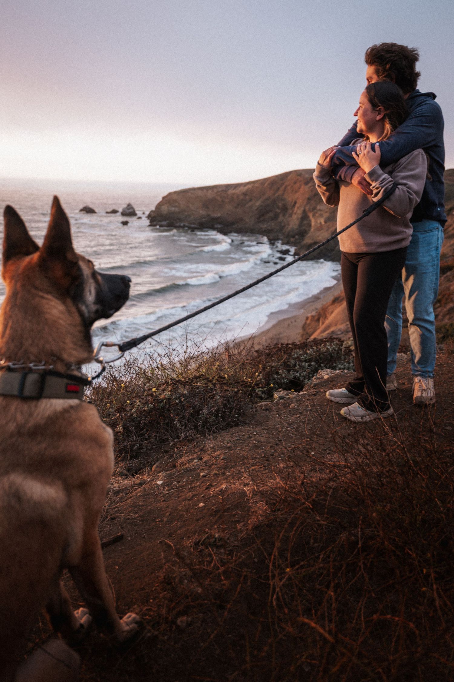 Couple embracing on coastal cliff at sunset with leashed German Shepherd dog overlooking rocky Pacific Ocean beach.