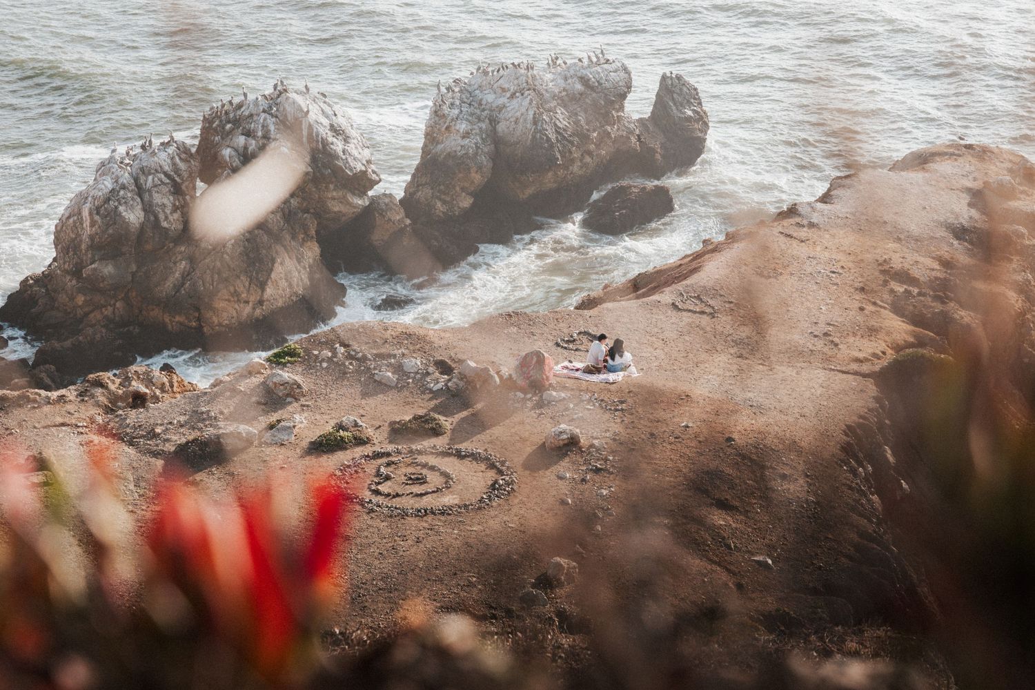 Aerial view of a rocky coastline with ocean waves, sandy shore, and a spiral rock art design near the water's edge.