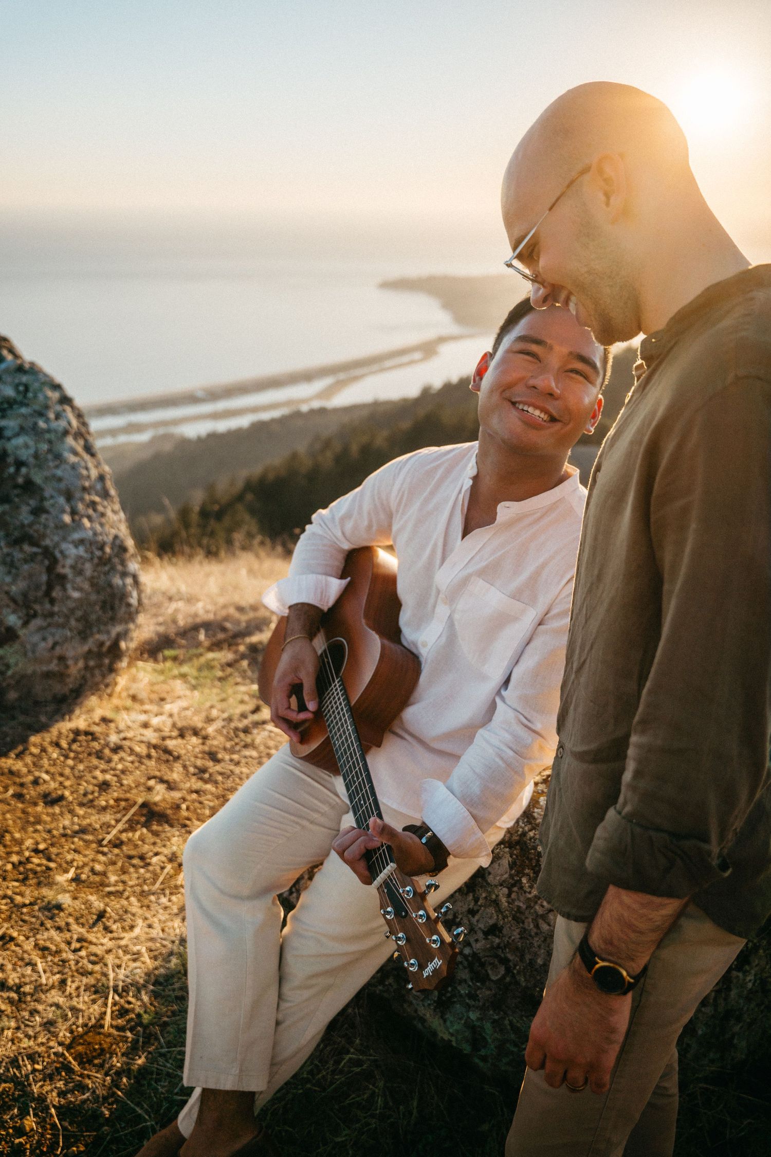 Two men share a romantic sunset moment on a hilltop, one playing acoustic guitar with ocean views behind them.