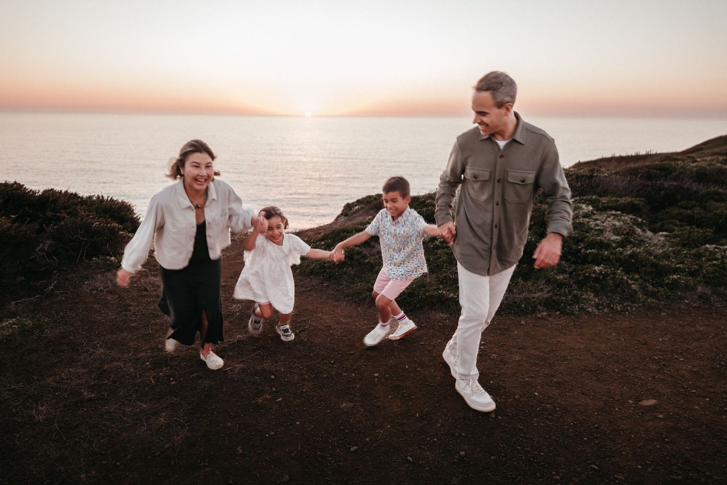 Family of four holding hands and running along a coastal cliff path at sunset with ocean views.