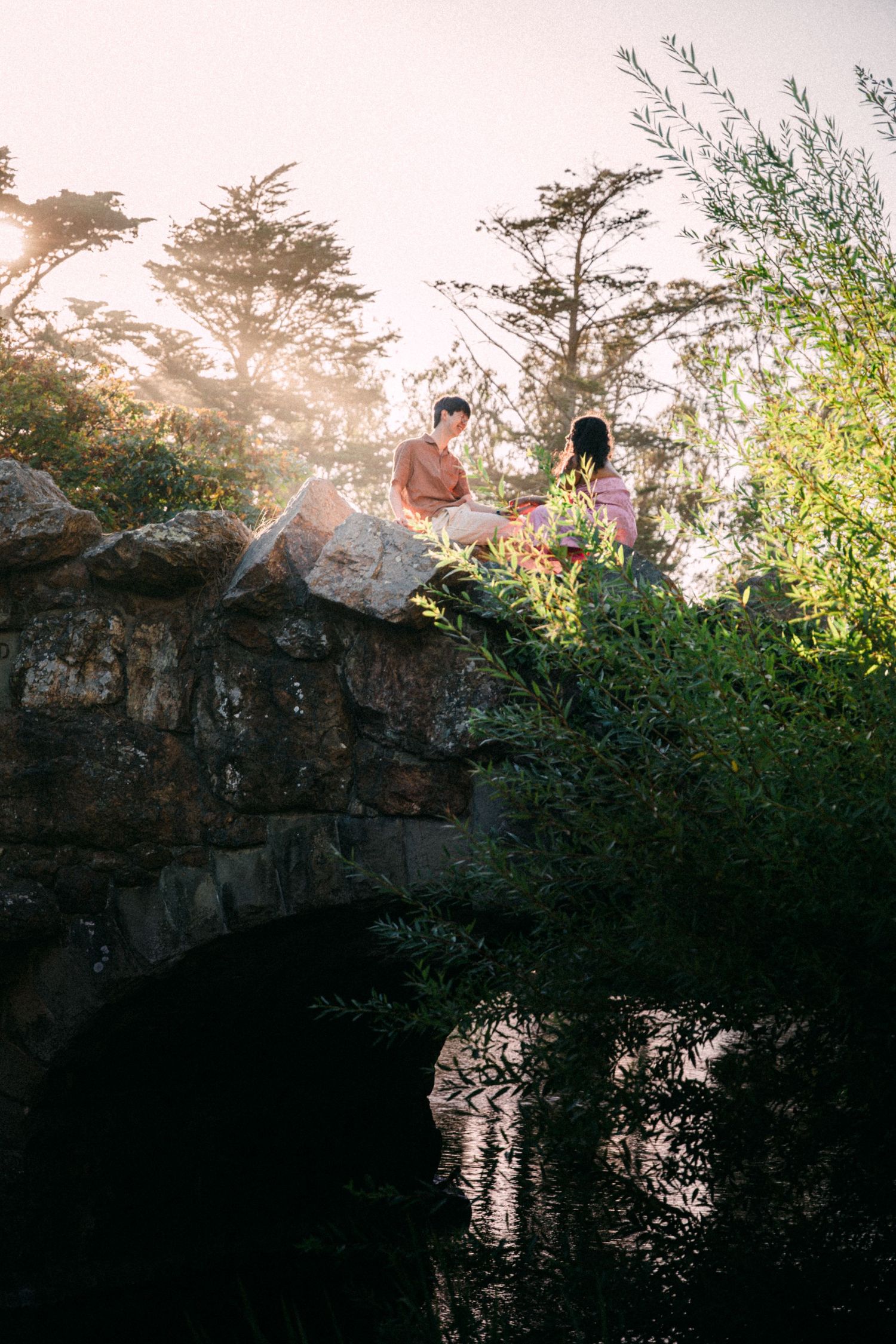 Couple sitting on a stone bridge surrounded by lush greenery and trees with soft golden sunlight filtering through the mist.