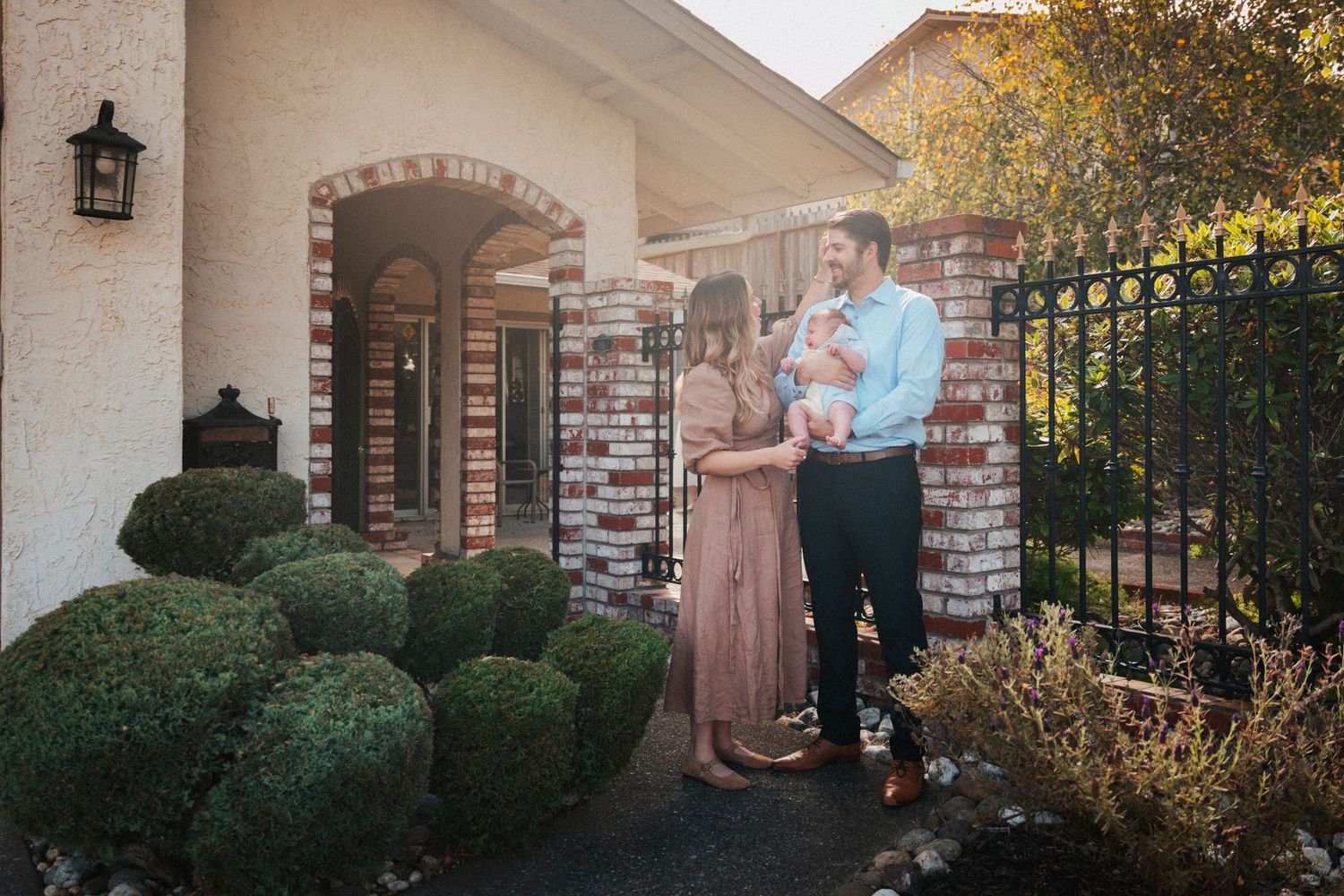 Young family with newborn baby poses outside brick home entrance with iron gate and manicured shrubs.