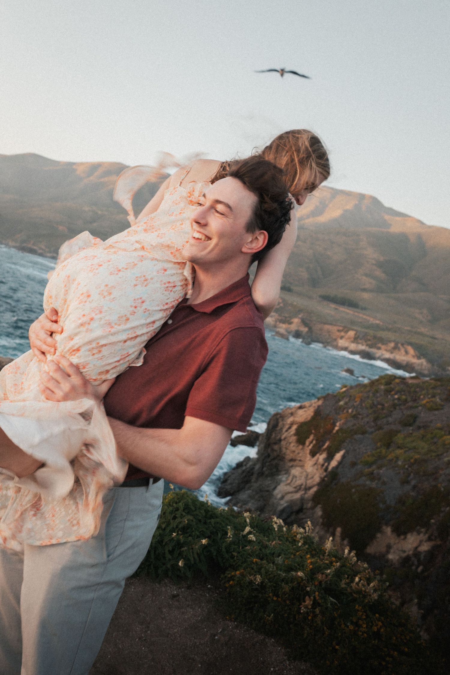 Happy man lifting woman in his arms at a scenic coastal overlook during golden hour with mountains in background.