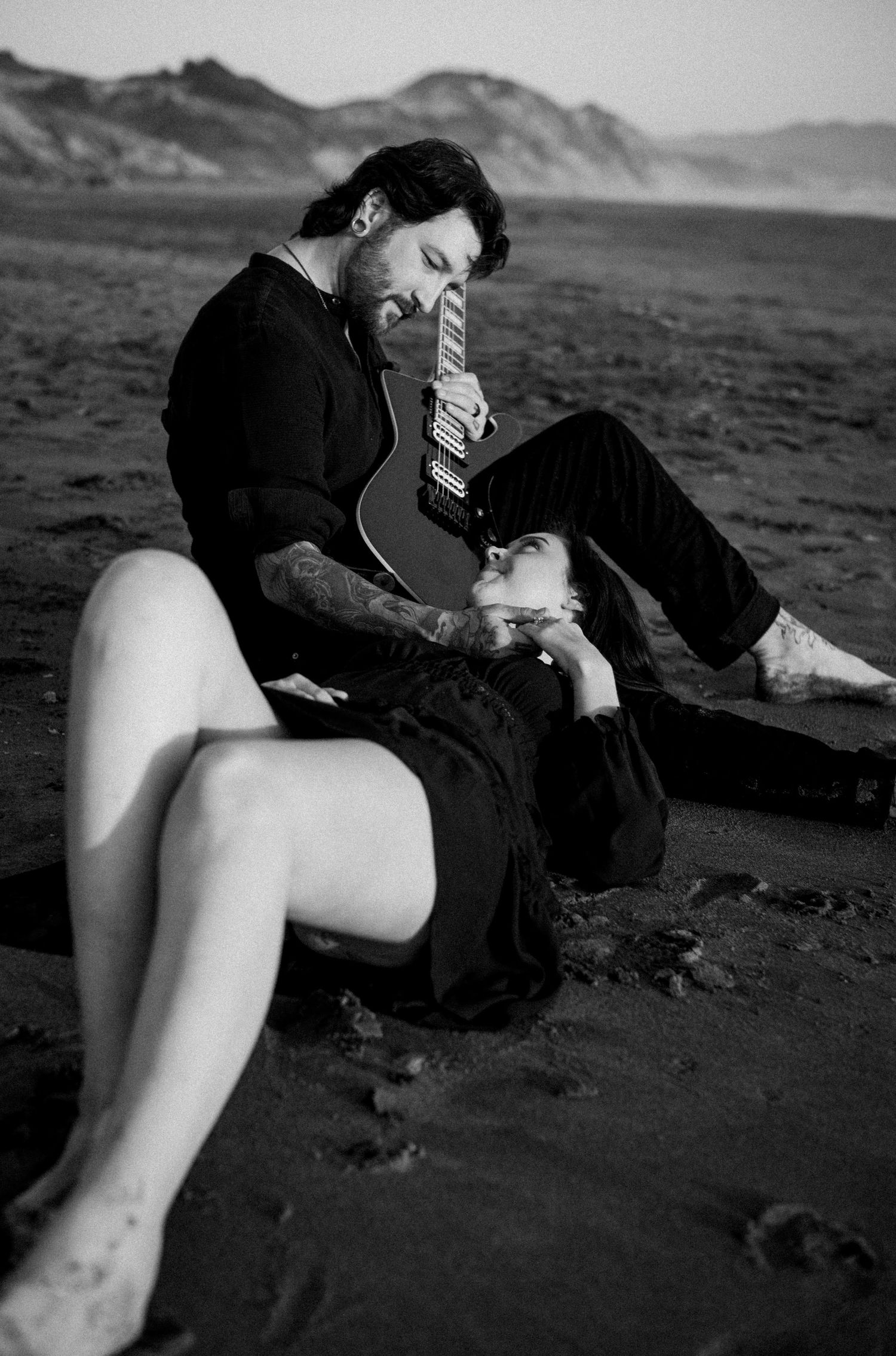 Black and white photo of a tattooed couple sitting on a beach, man holding guitar while woman rests her head back.