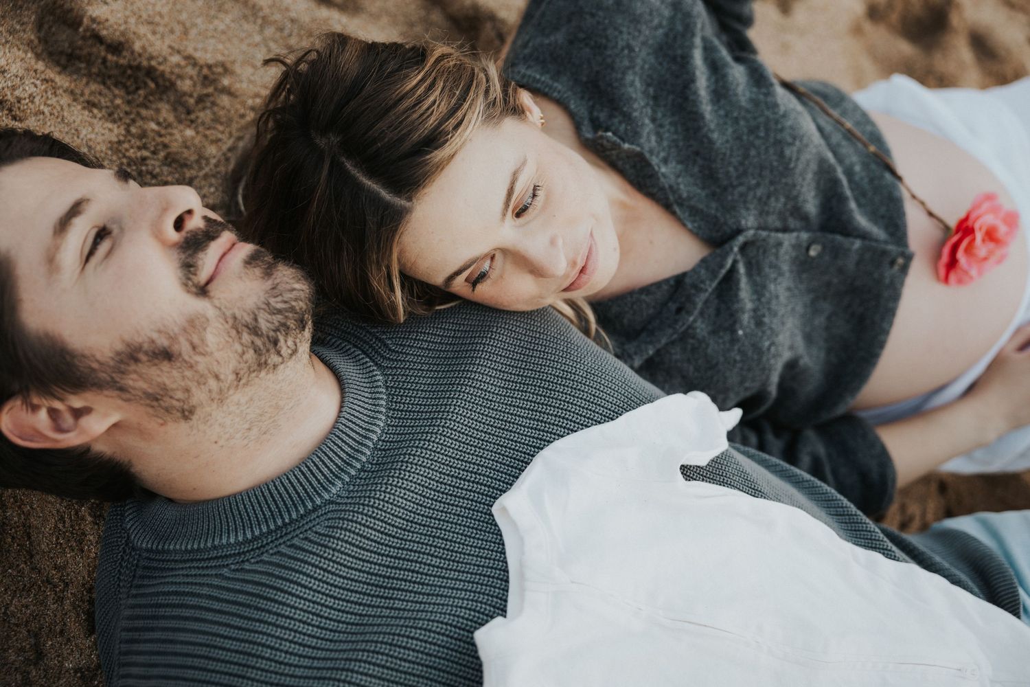 A couple lying on sandy ground, gazing at each other tenderly, the woman wearing a gray sweater with a pink flower.