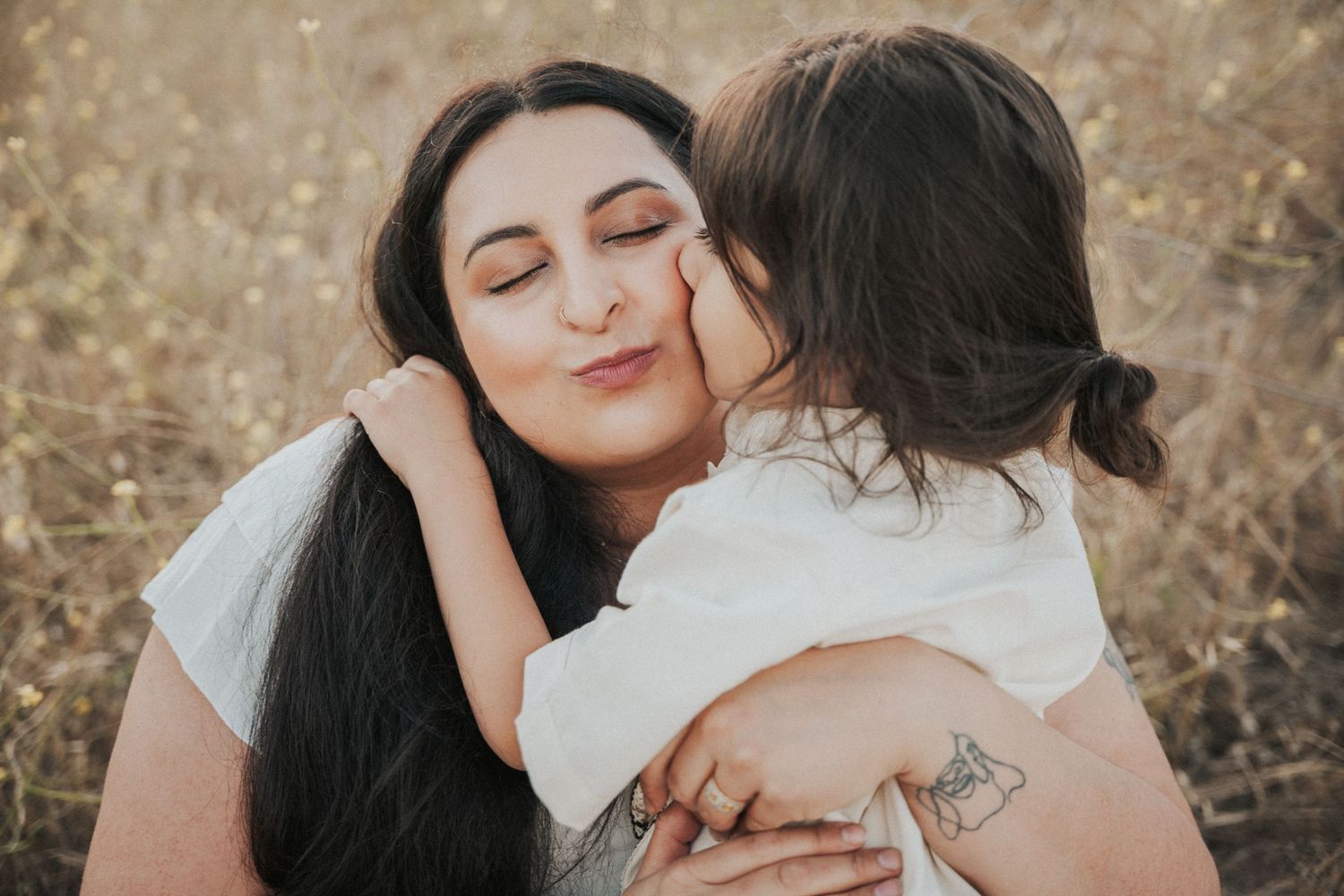 A mother with long dark hair receives a sweet kiss on the cheek from her young child outdoors.