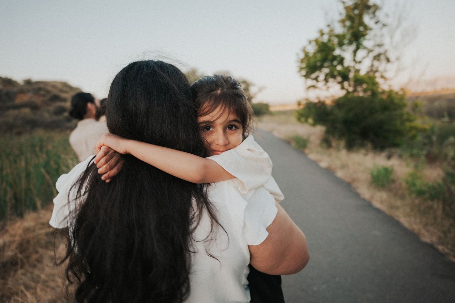 A young girl hugs a dark-haired woman on a scenic path, smiling warmly in a heartfelt outdoor embrace.