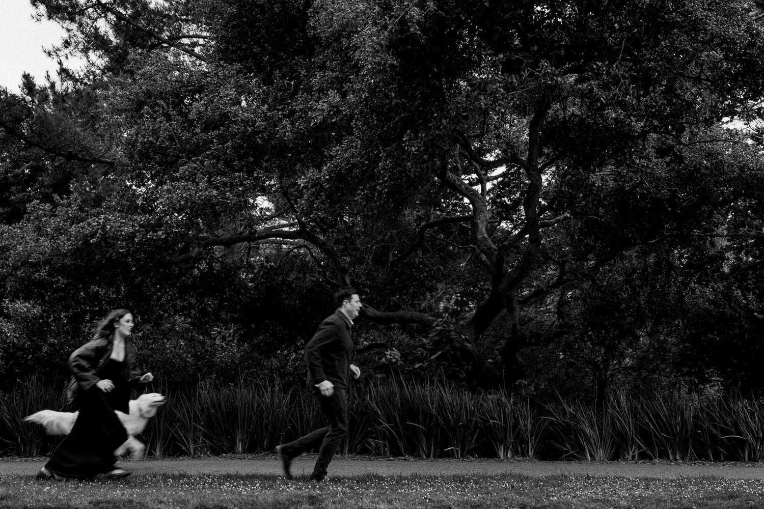 Black and white photo of a couple running playfully through a lush park with tall trees in the background.Photography by The Y.K. Studio.