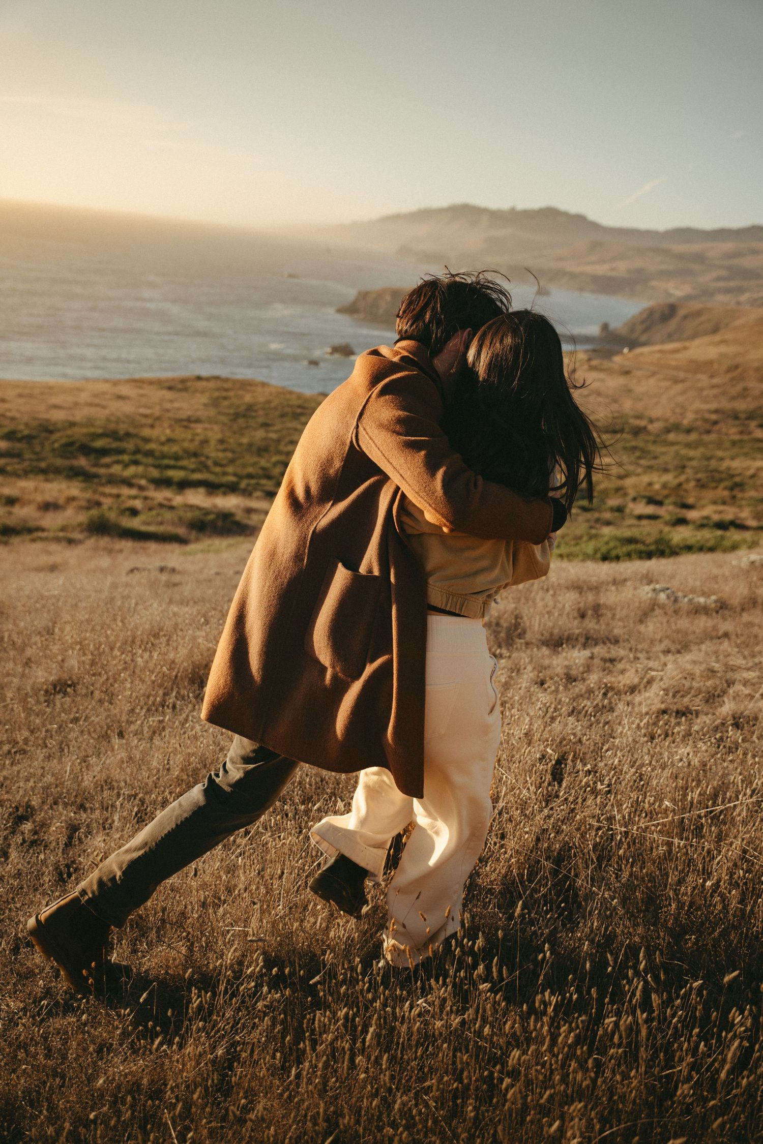 A couple in a tan coat and cream pants embrace and twirl on a grassy coastal hillside overlooking the ocean at golden hour.