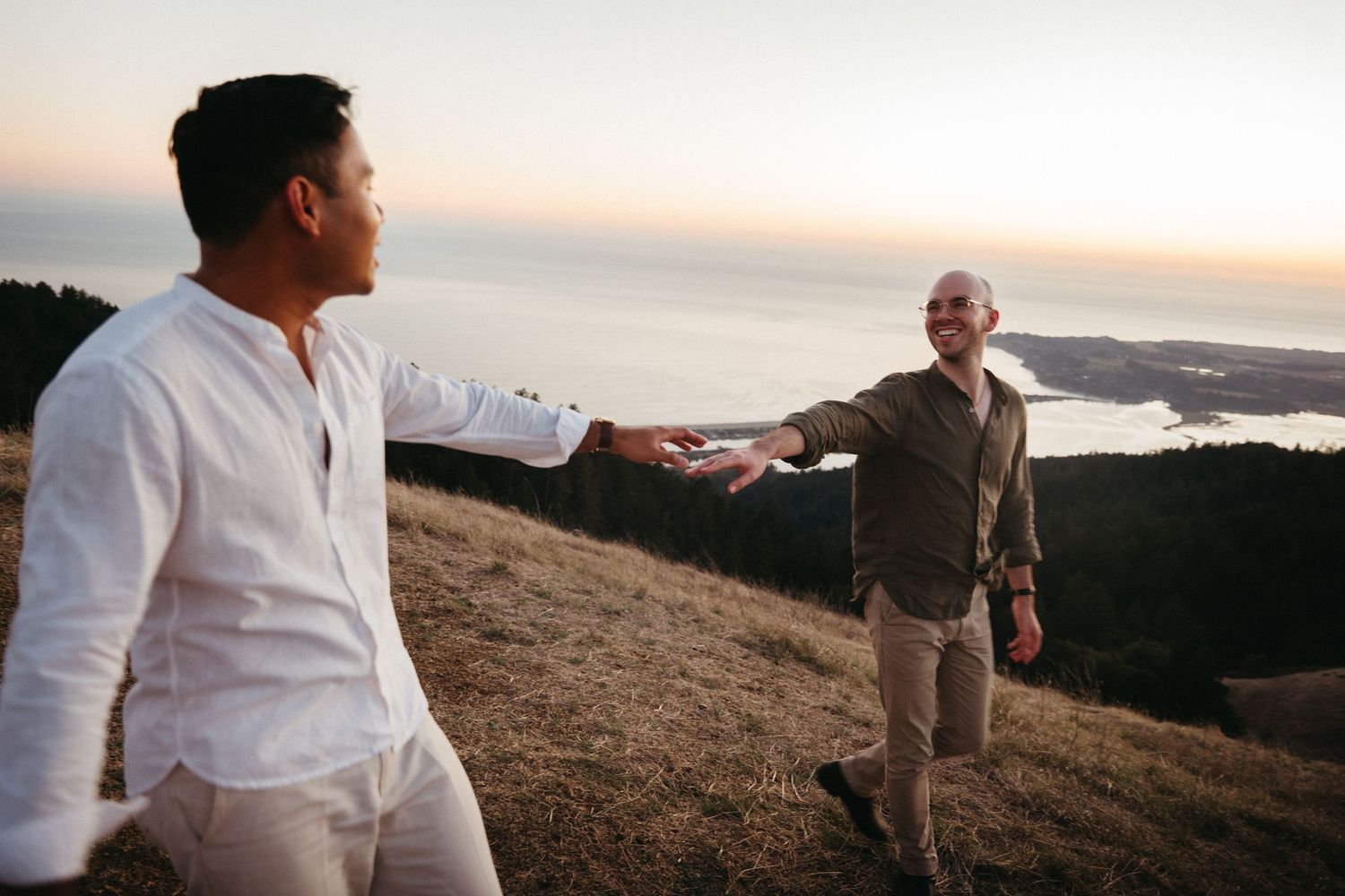 Two men reaching toward each other on a scenic hilltop at sunset with ocean views in the background.
