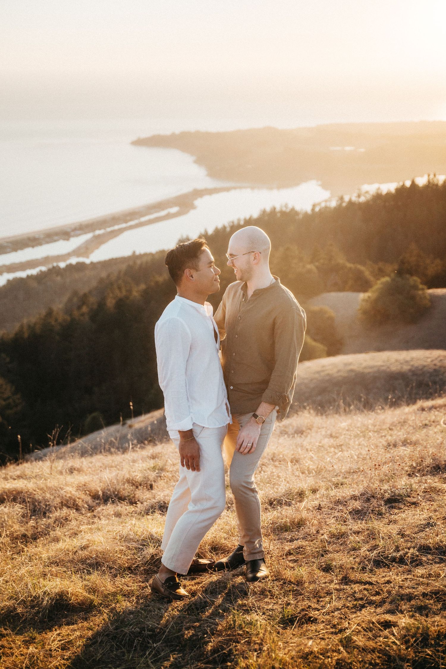 Two men share a romantic moment on a golden hillside at sunset with a scenic coastal landscape behind them.