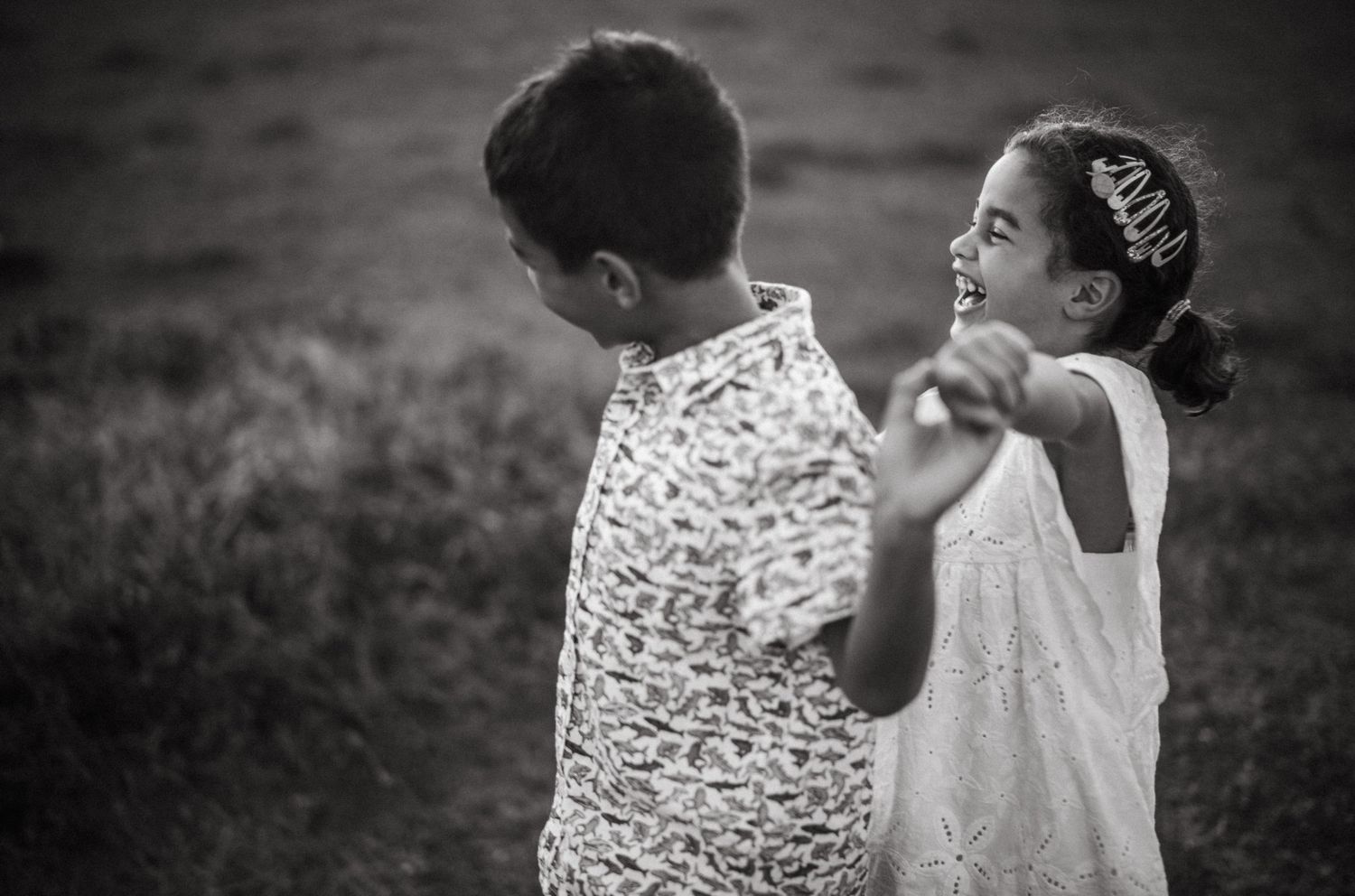 Two children dance and laugh together outdoors in a black and white photo, girl in white dress, boy in patterned shirt.
