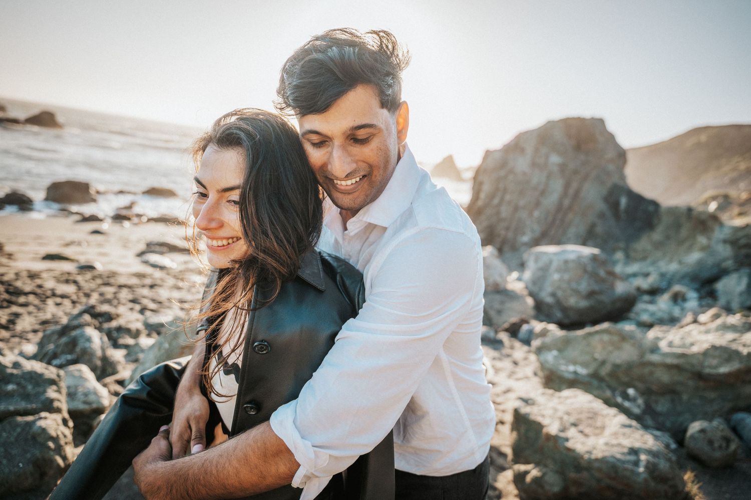 Man in white shirt embracing smiling woman on rocky beach at golden hour, romantic couples photography.