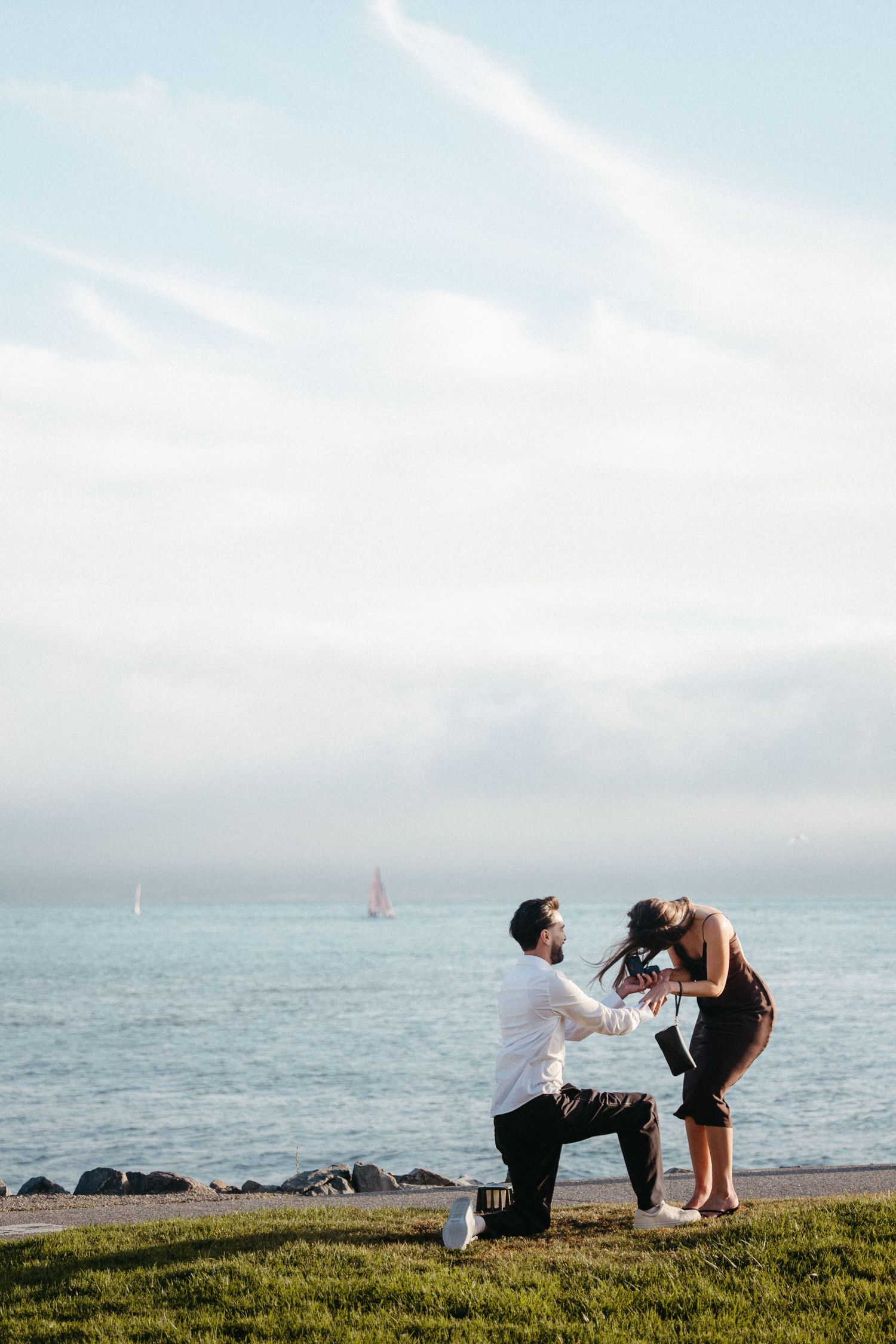 Man proposes on one knee to woman by the ocean shore on a cloudy day near sailboats.