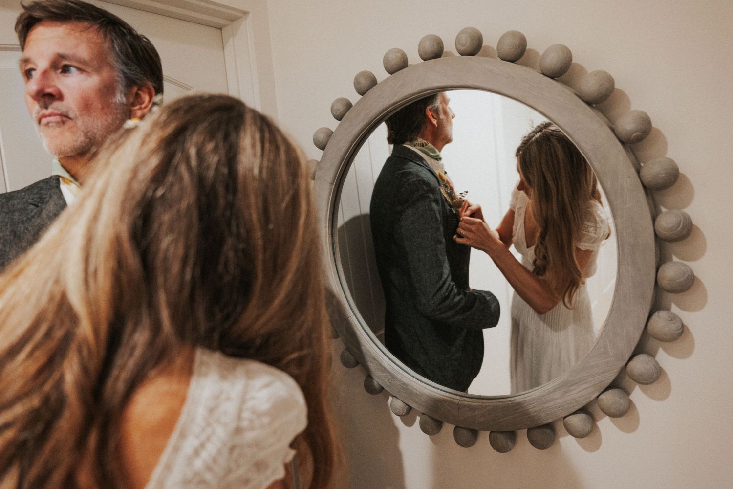 Couple sharing an intimate moment reflected in a decorative round mirror during wedding preparations.