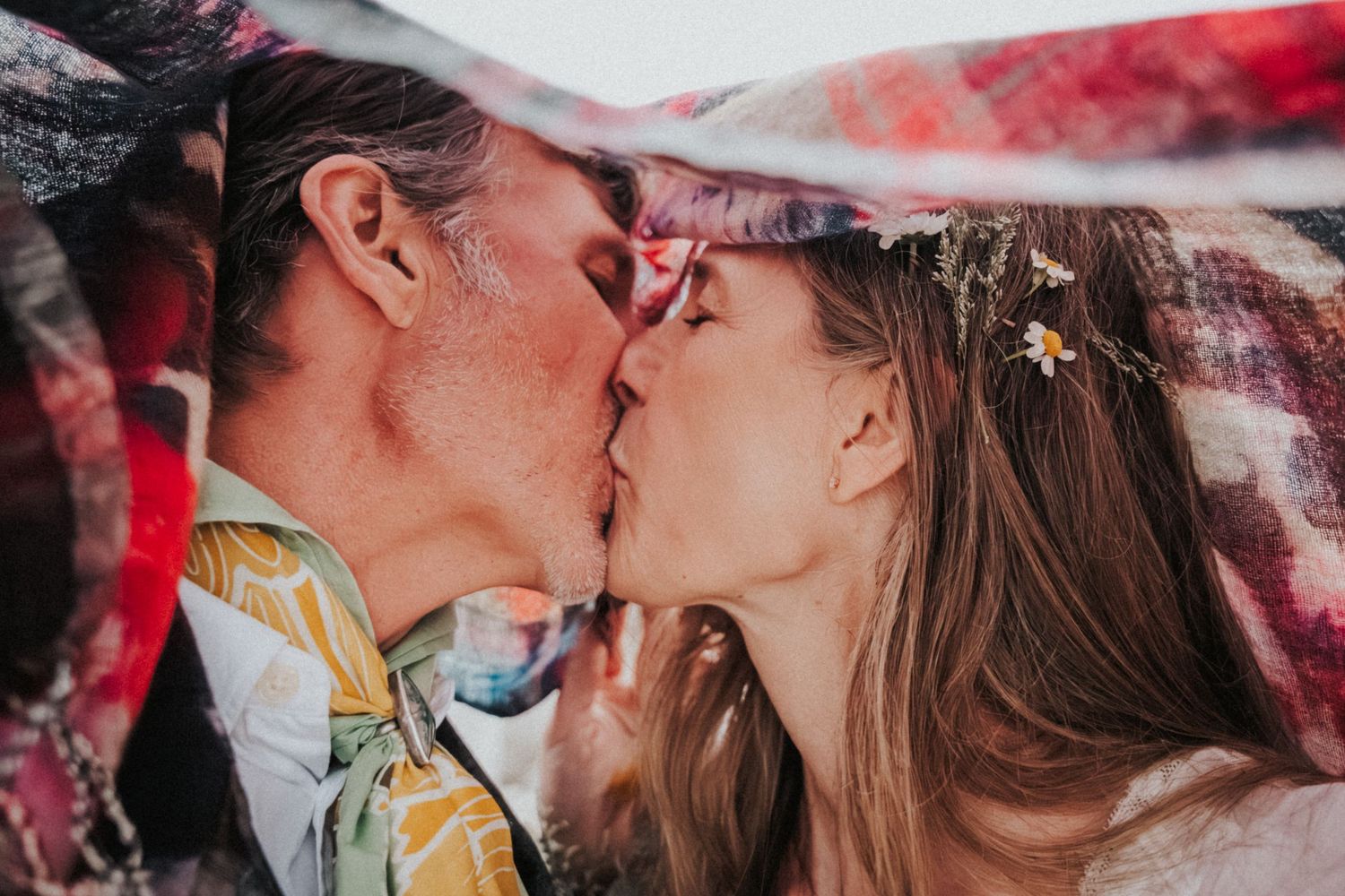 A couple shares a romantic kiss under a colorful patterned blanket at their wedding.