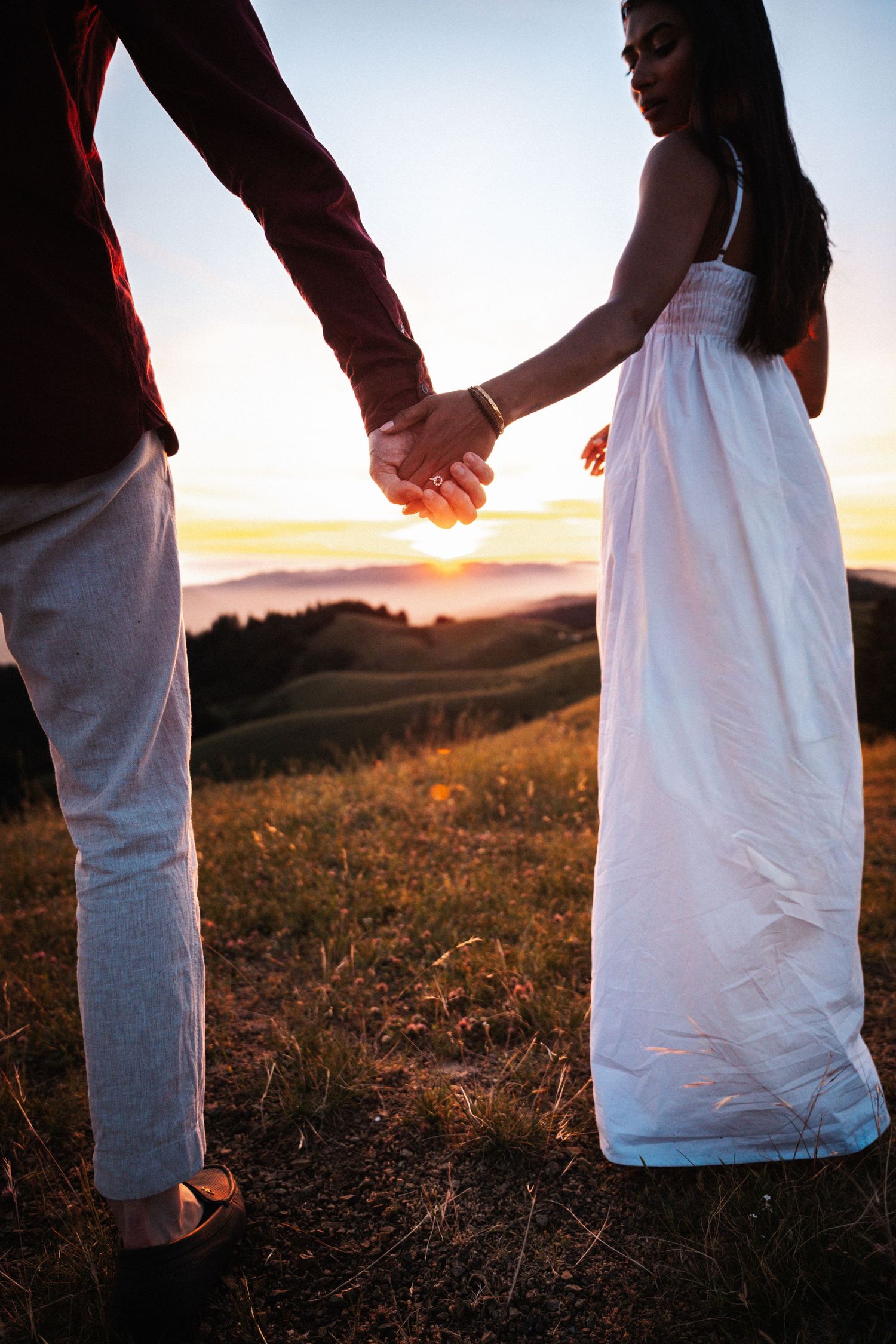 Couple holding hands at sunset on a grassy hill, woman in white dress, golden sky backdrop.