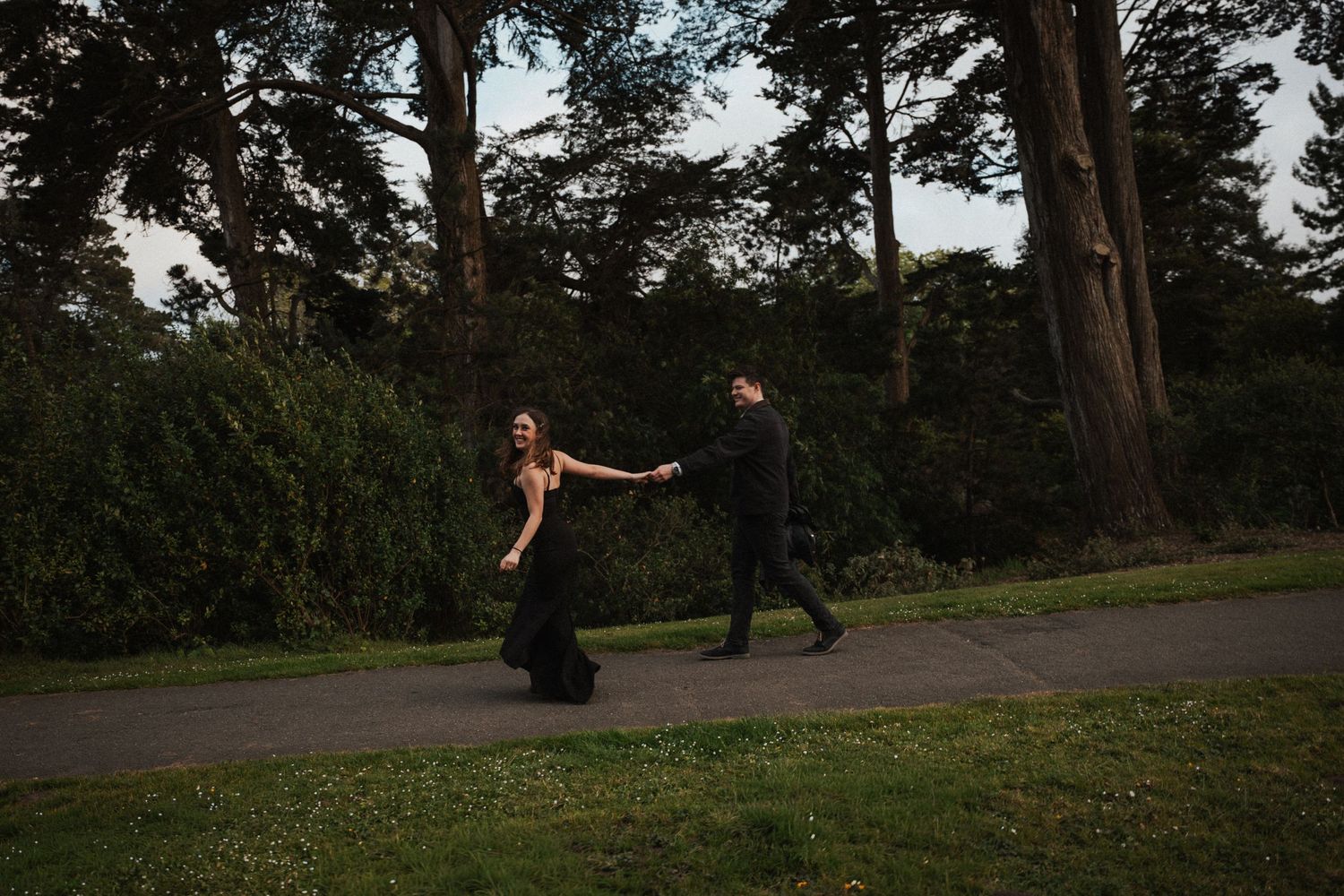 Couple in black formal attire holding hands walking on a path surrounded by lush green trees at dusk.