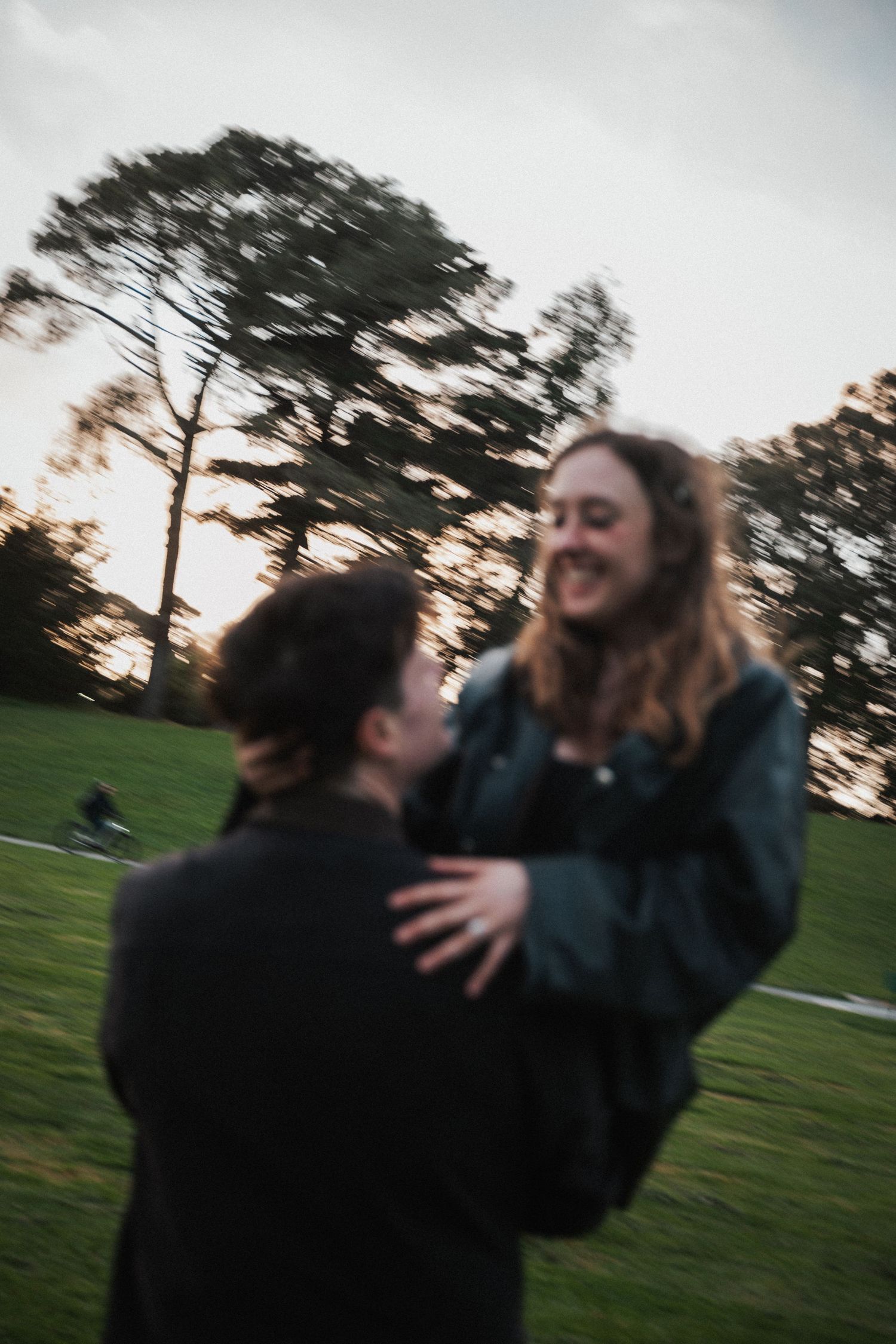 A man lifts a smiling woman in a park at dusk, both wearing dark jackets in a joyful embrace.
