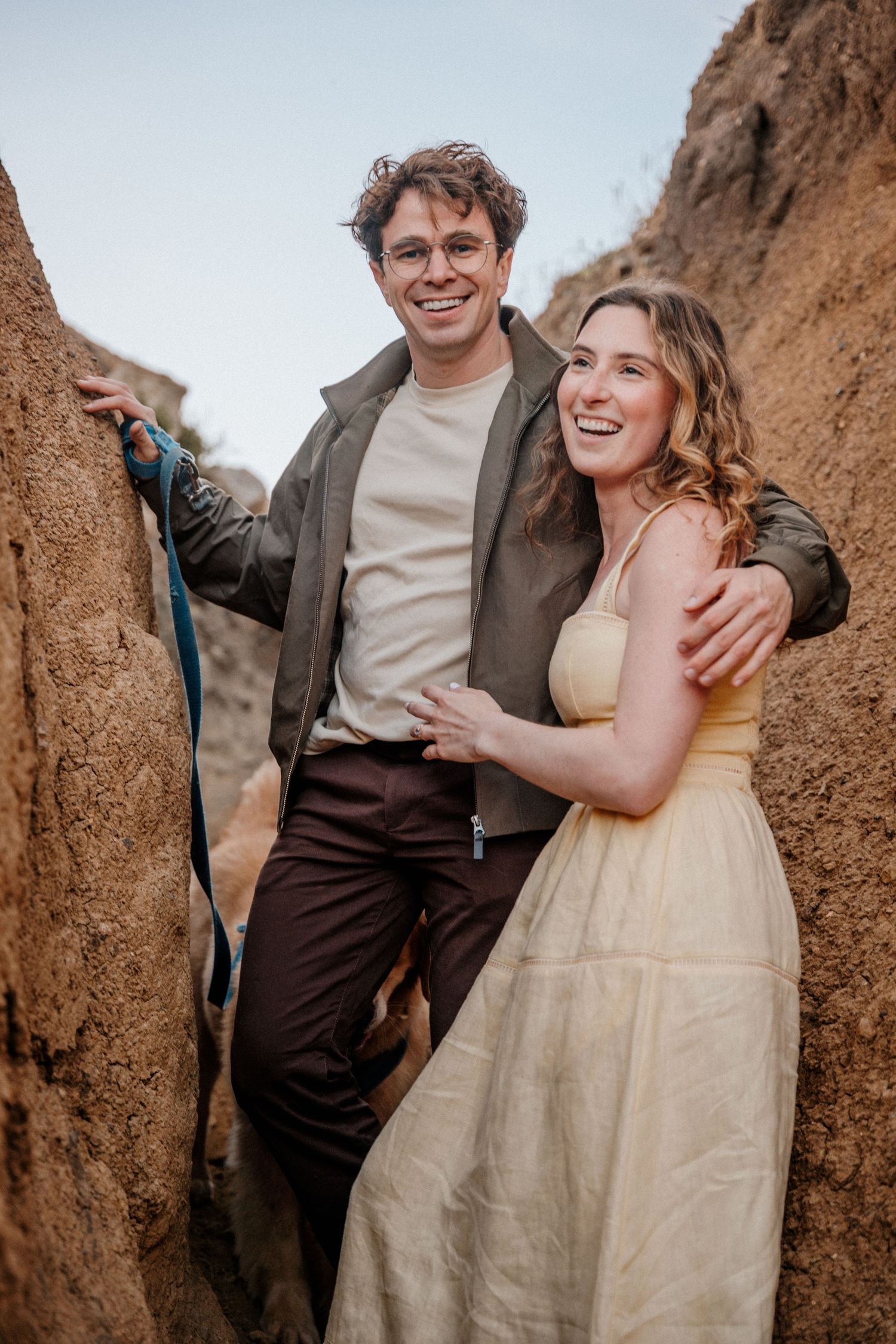 Smiling couple posing between rocky formations, man in glasses and vest, woman in yellow dress.