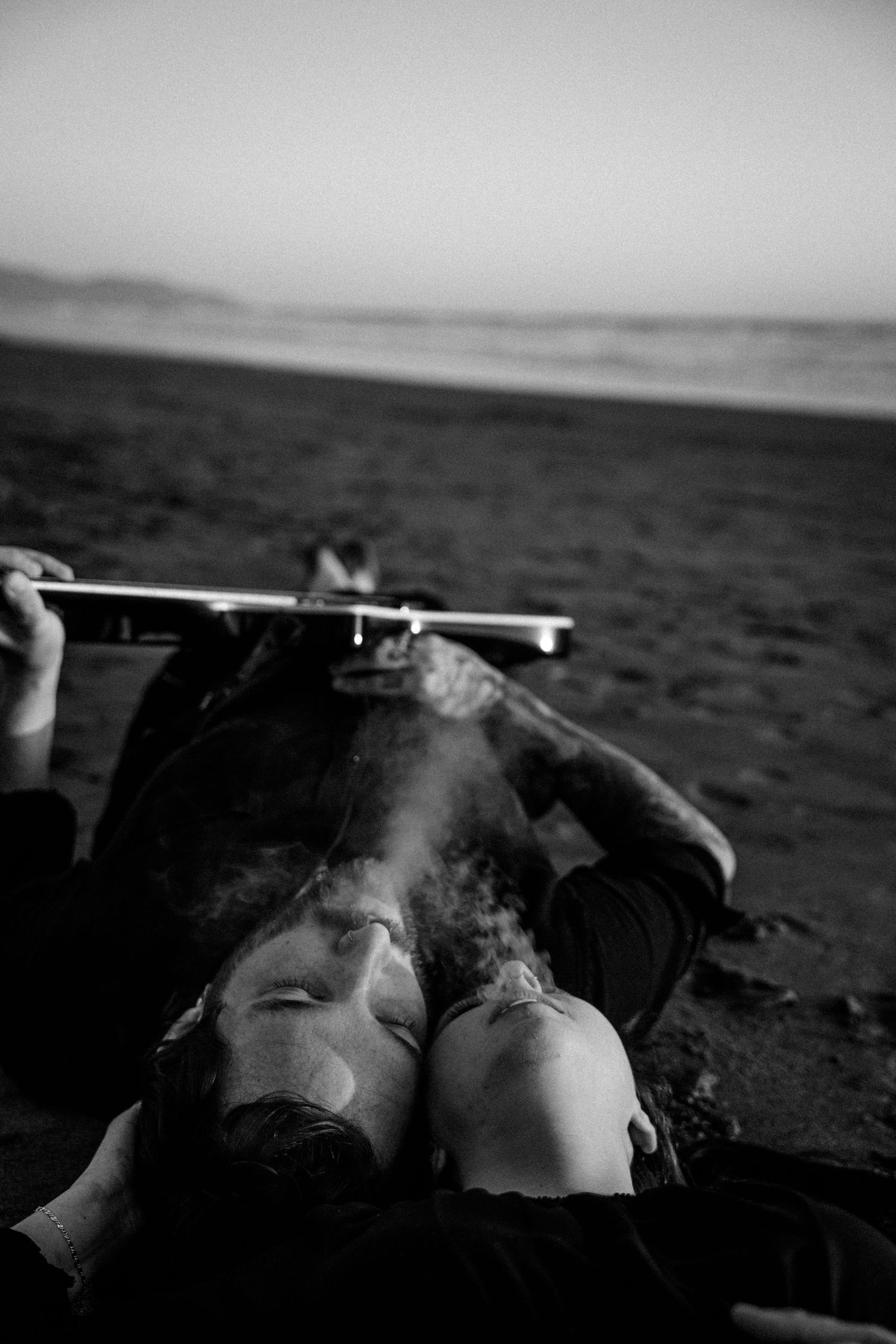 Black and white photo of couple lying on beach, sharing a kiss with smoke rising between them.