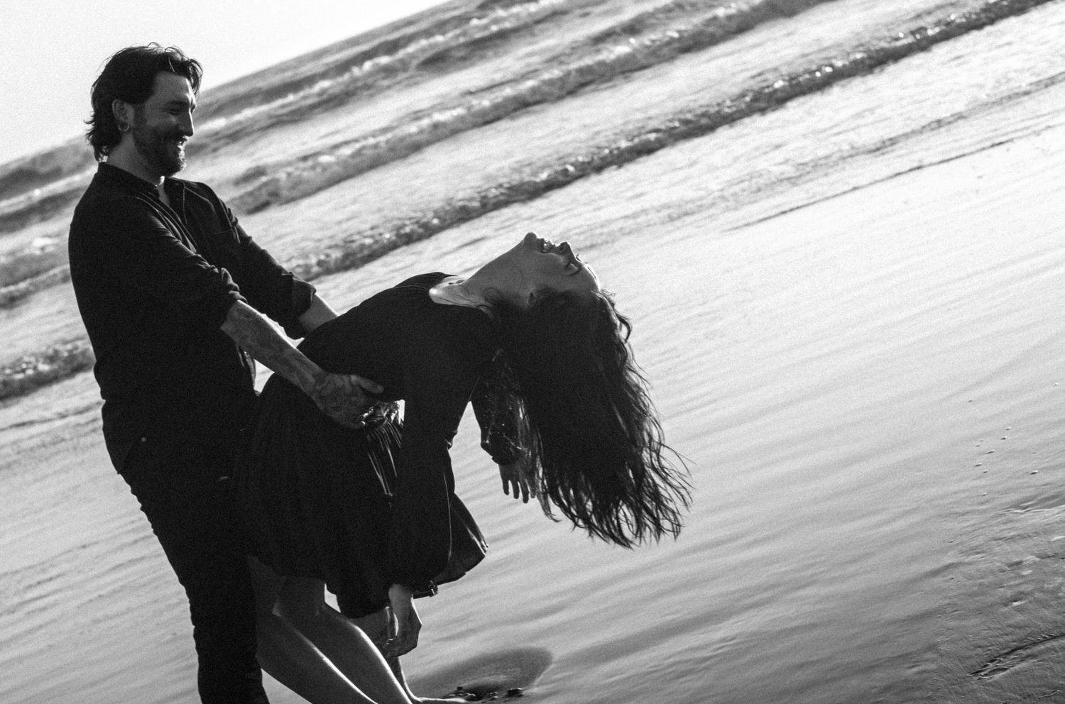 Man lifting woman in his arms at beach shoreline in dramatic black and white photograph.