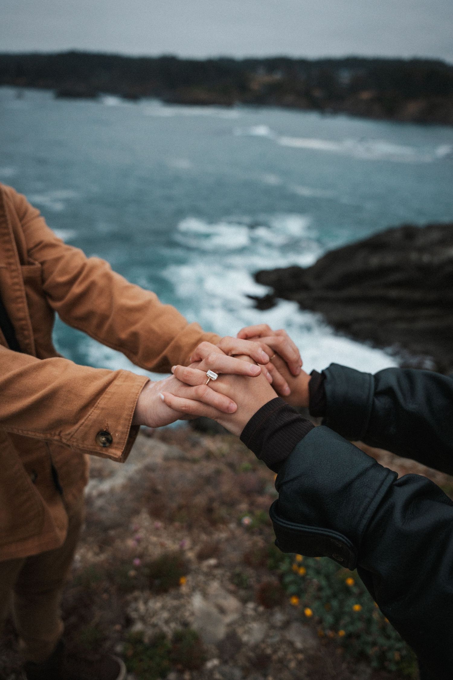 Couple holding hands with engagement ring overlooking rocky ocean coastline.