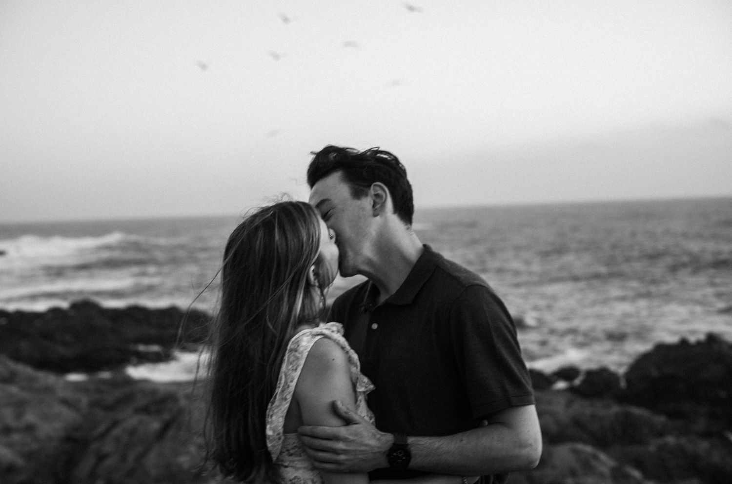 Black and white photo of a romantic couple embracing near rocky ocean shore with birds flying in the background.