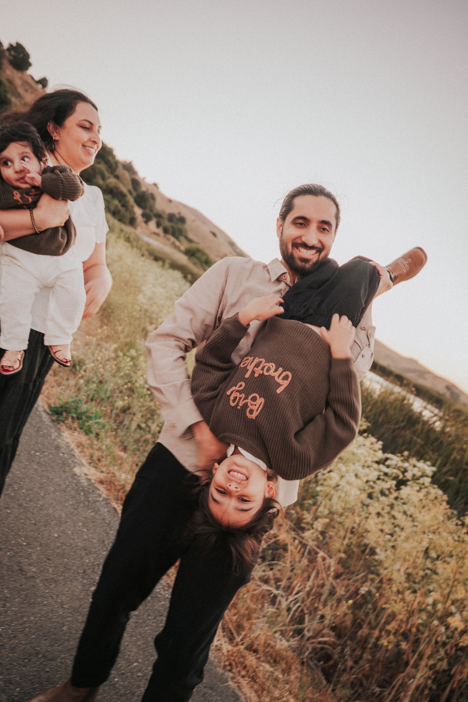 Joyful family photo with father holding child upside down while mother holds baby.
